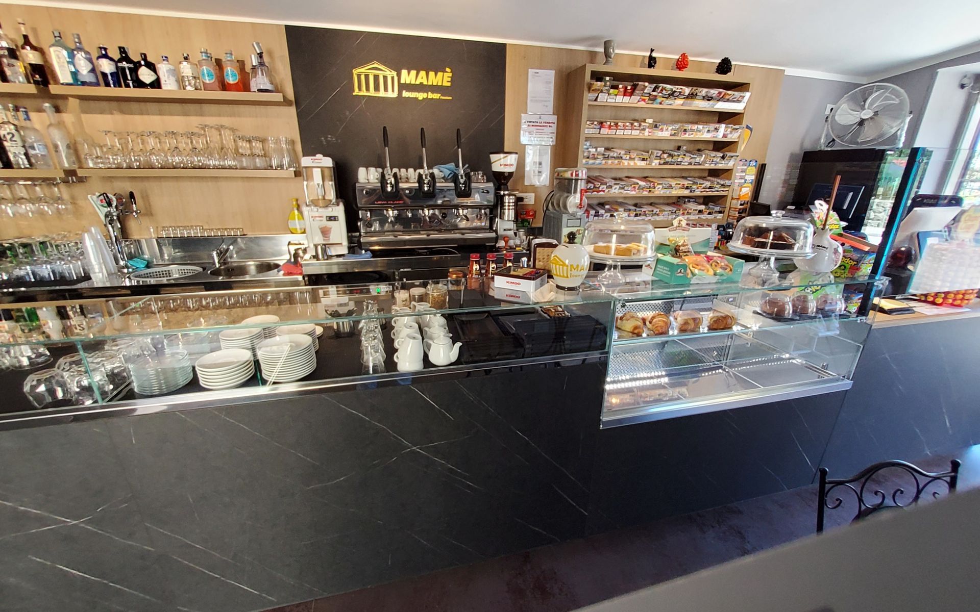 Vista interna di un bar con bancone che espone pasticcini, macchine per il caffè e scaffali pieni di bicchieri e bevande.