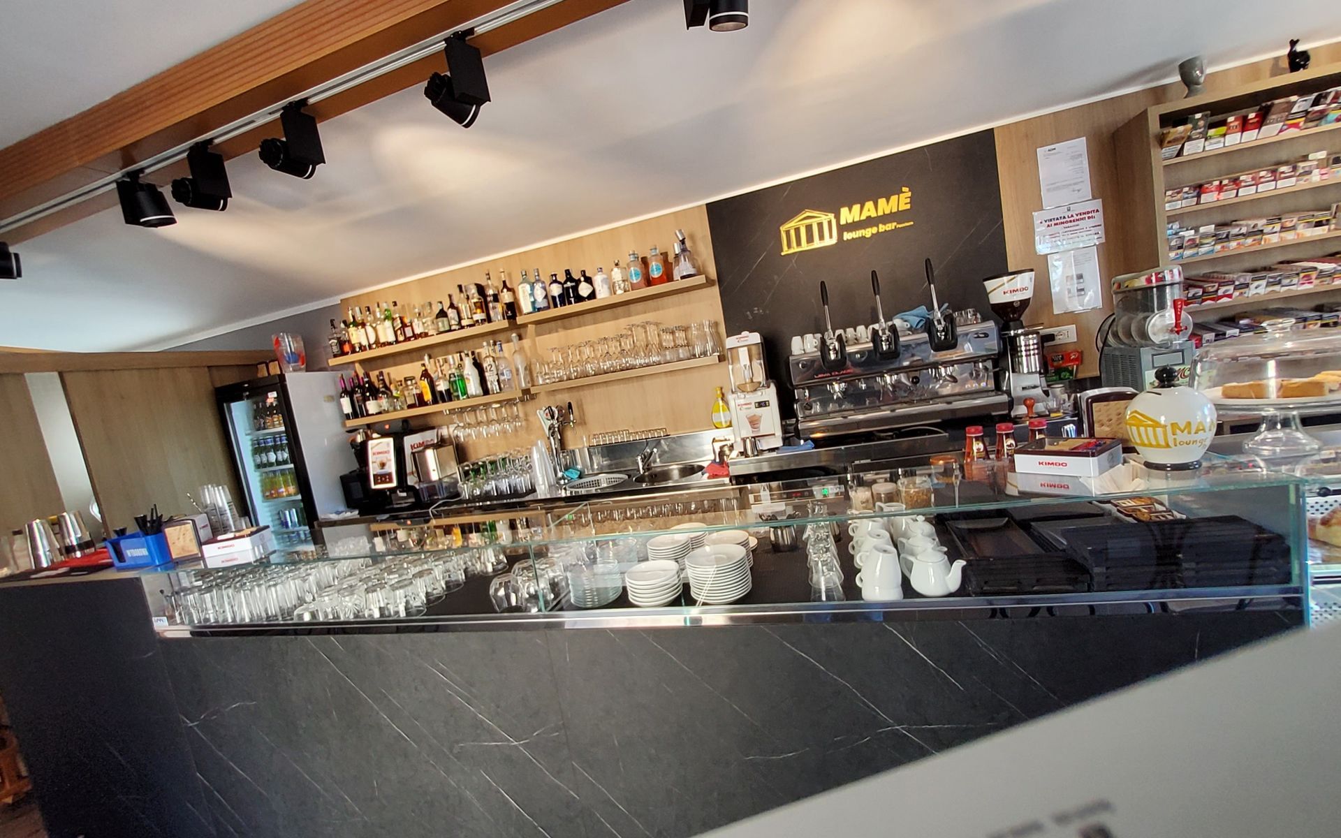 Vista interna di un bar con bancone scuro e bottiglie di liquore su uno scaffale. Sono visibili le macchine del caffè.