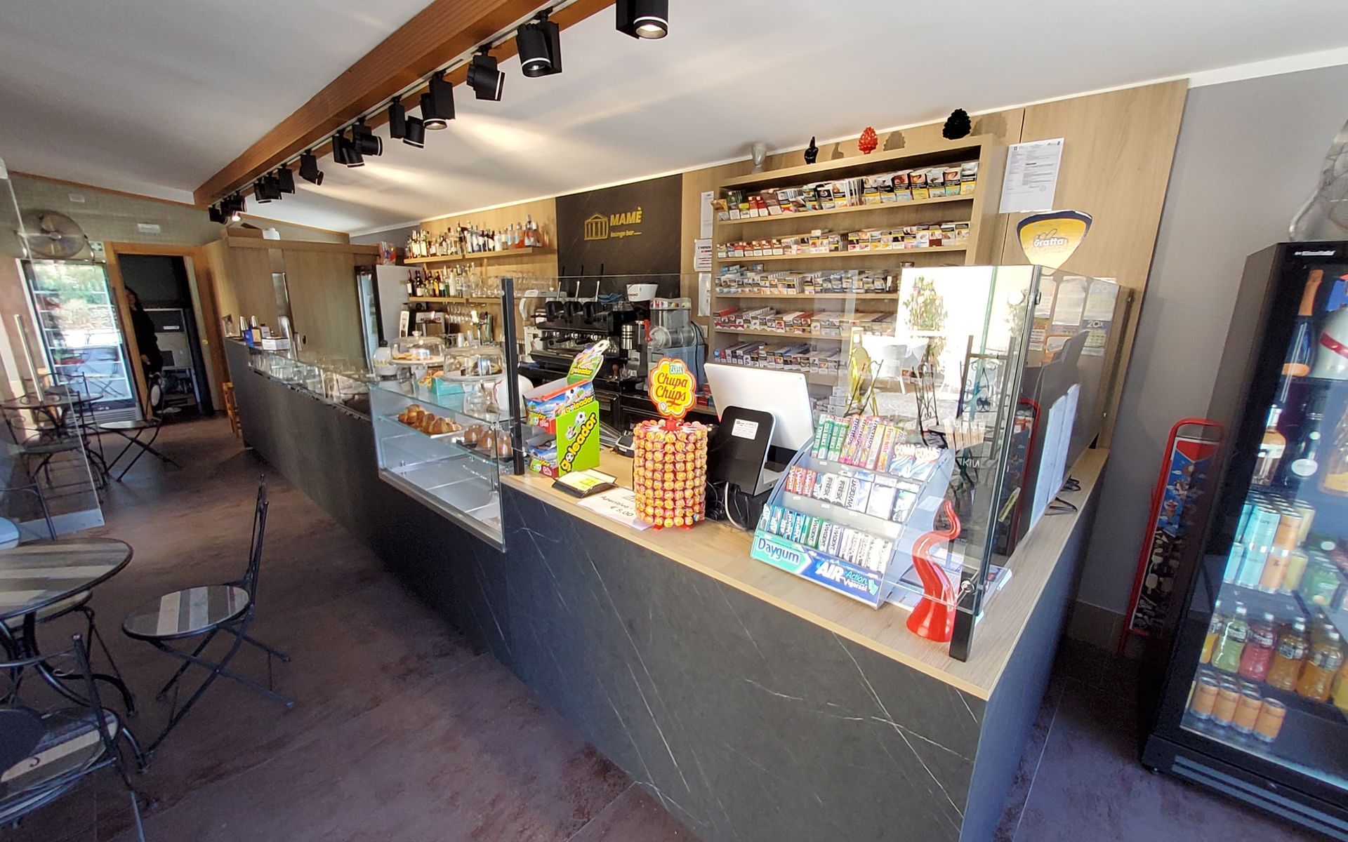 Interno di un bar con un lungo bancone che espone dolci e altri prodotti. Sono disponibili una macchina per il caffè, un frigorifero per le bevande e posti a sedere.