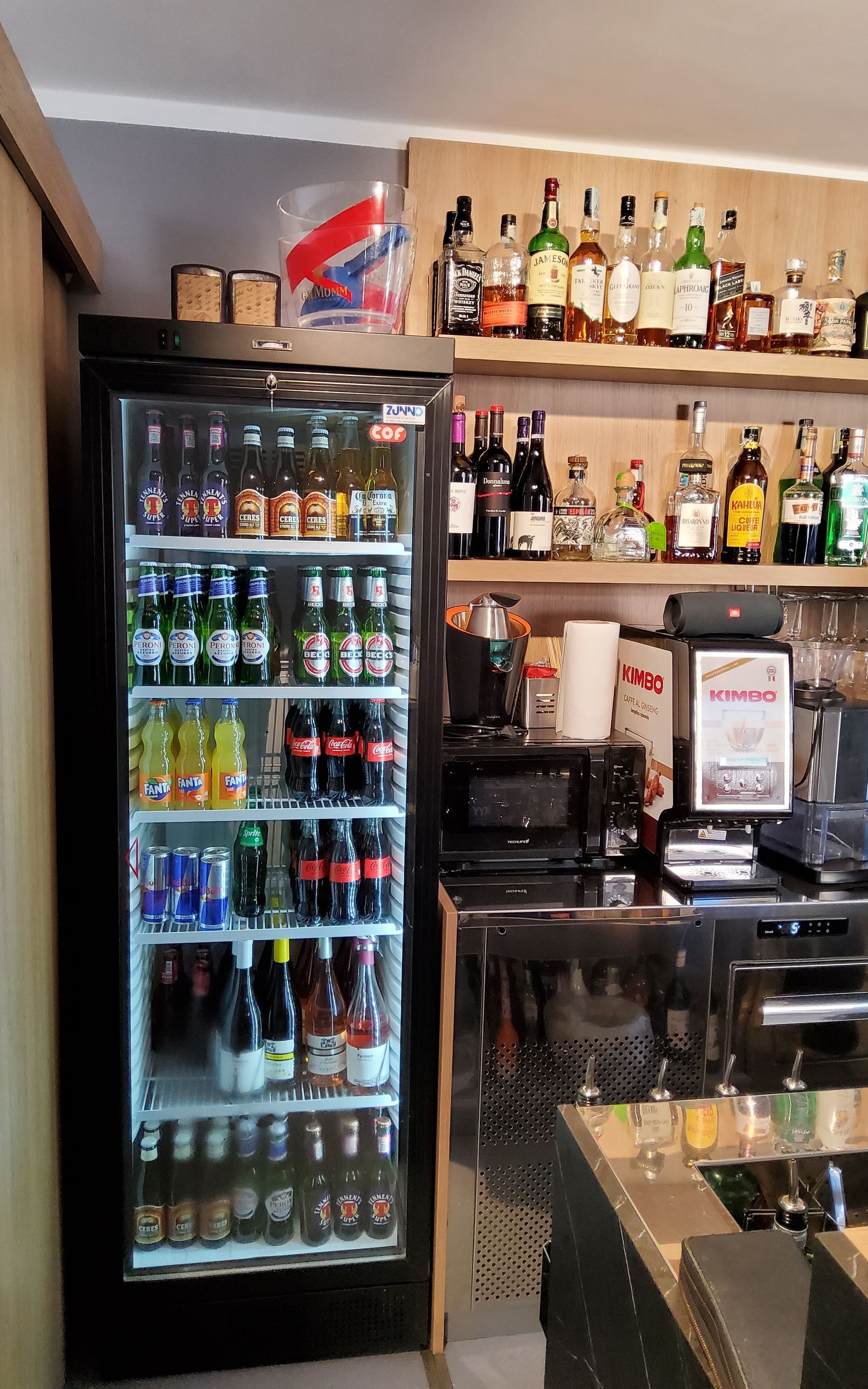 Frigorifero rifornito di bevande al bar, scaffali con bottiglie di liquore e un forno a microonde.