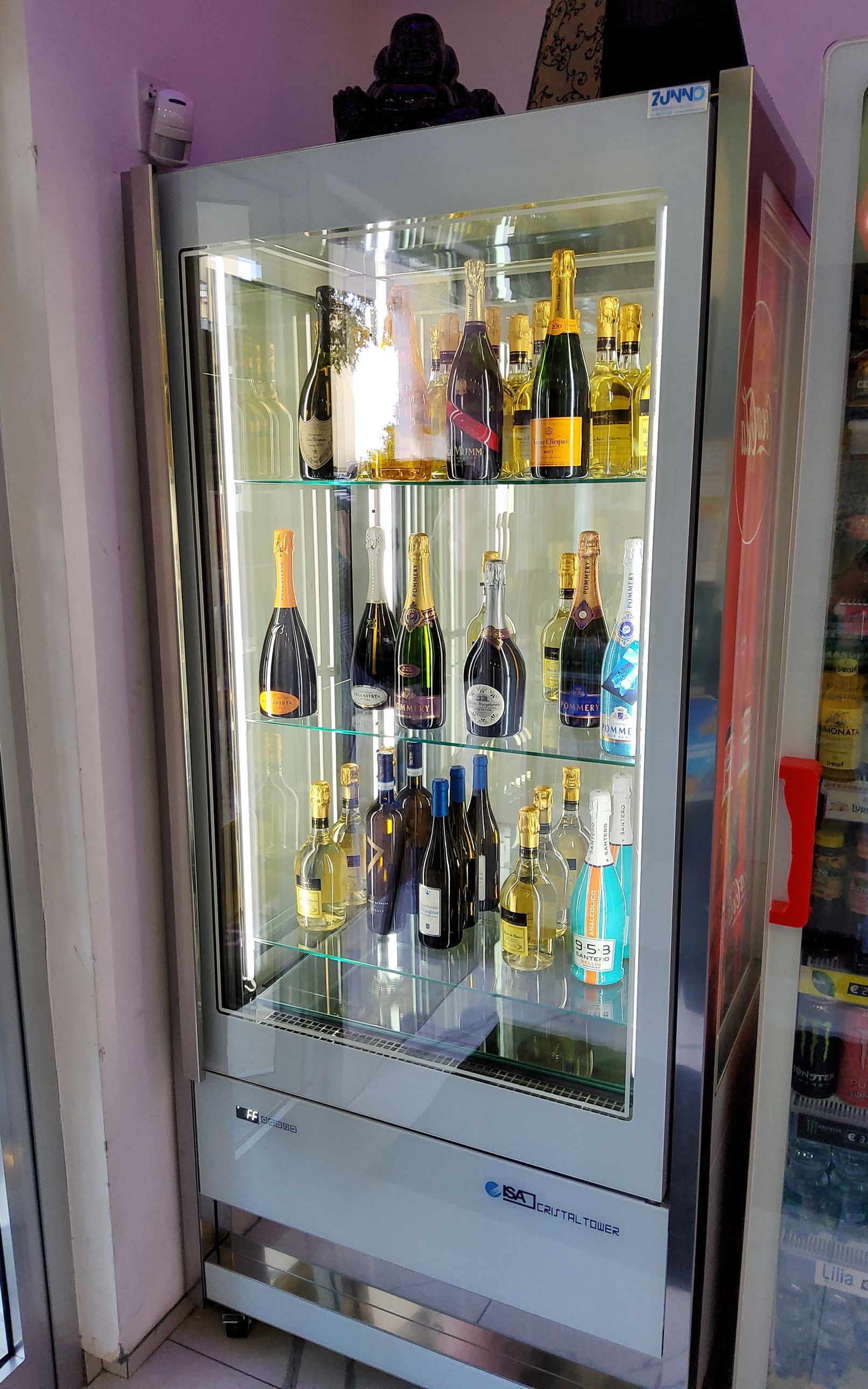 Vetrina con diverse bottiglie di alcolici. Ripiani illuminati su un interno bianco, maniglia della porta rossa.