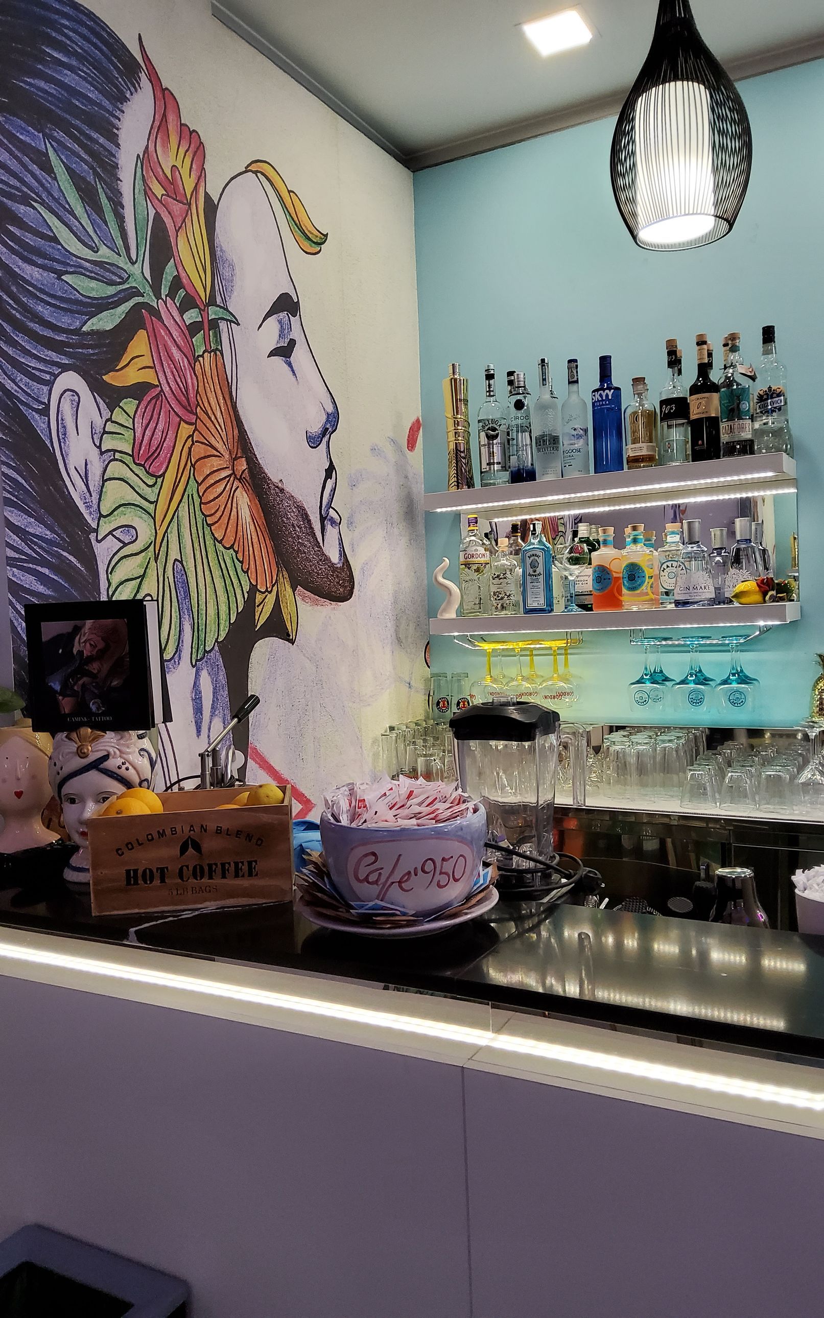 Bar con murale colorato, bottiglie sugli scaffali e bicchieri. Sul bancone sono presenti una torta e un'insegna in legno.