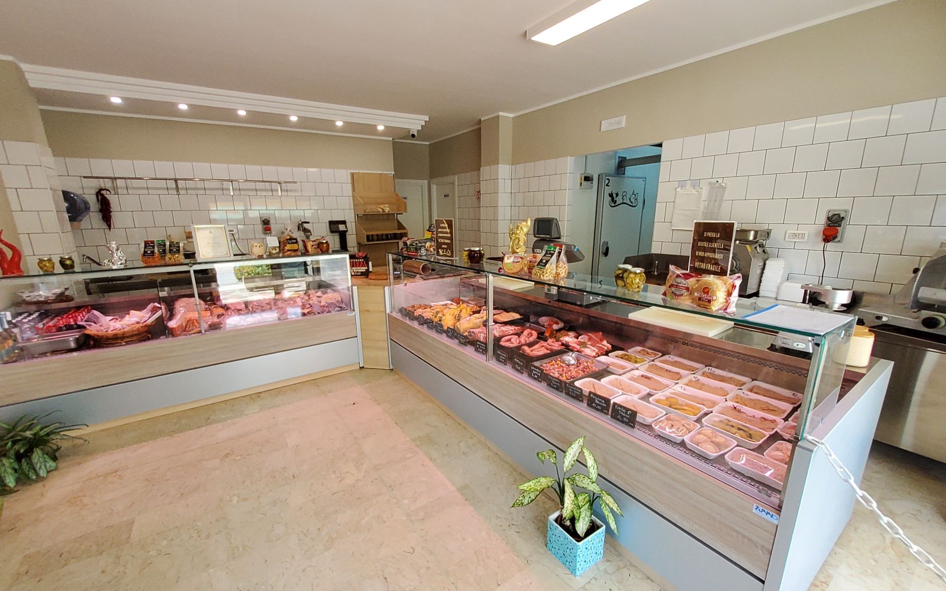 Interno di una macelleria con vetrine espositive di carni e prodotti confezionati. Pareti beige chiaro e illuminazione fluorescente.