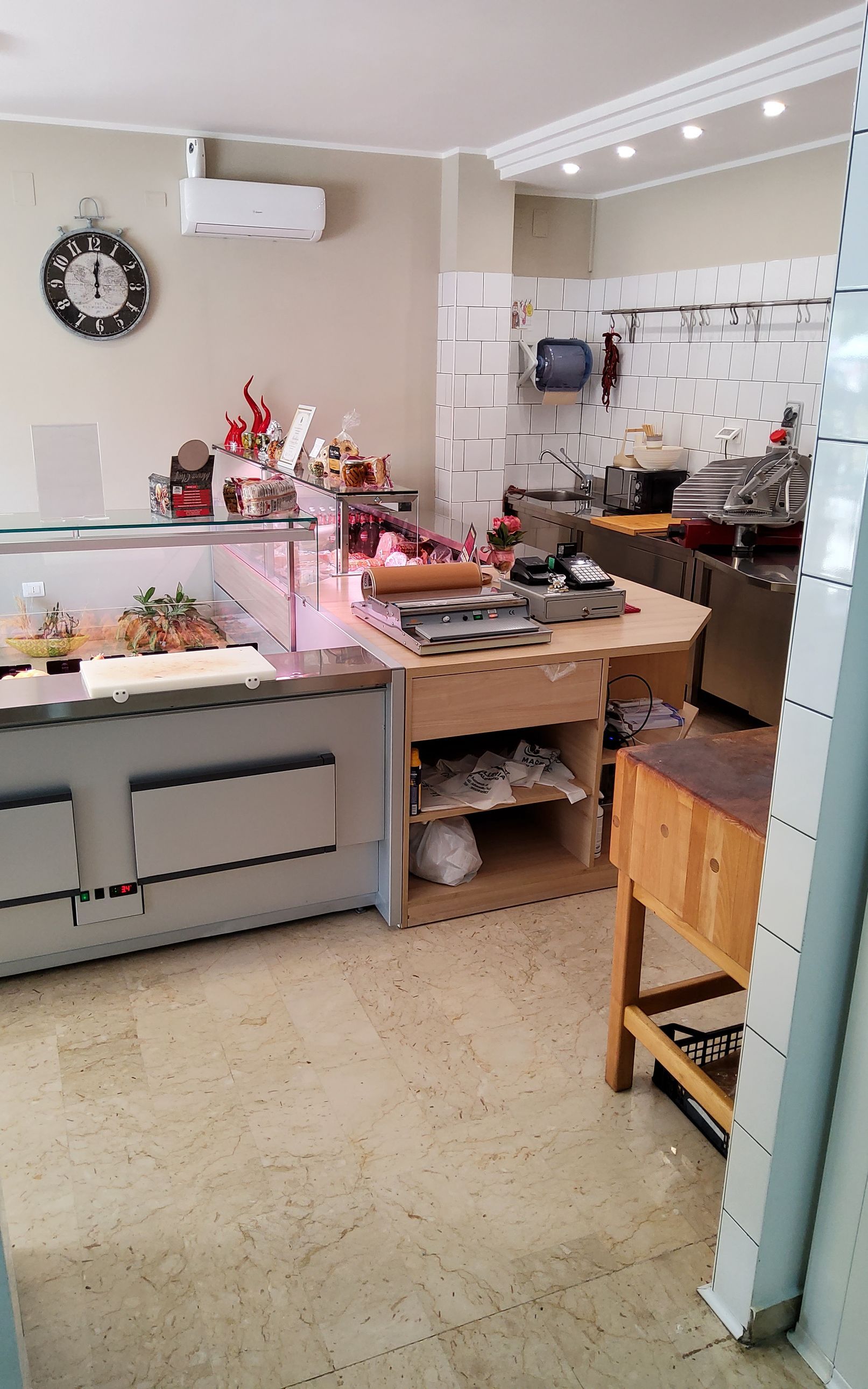 Interno di una macelleria con vetrina, bancone e attrezzature. Pavimento in piastrelle beige chiaro.