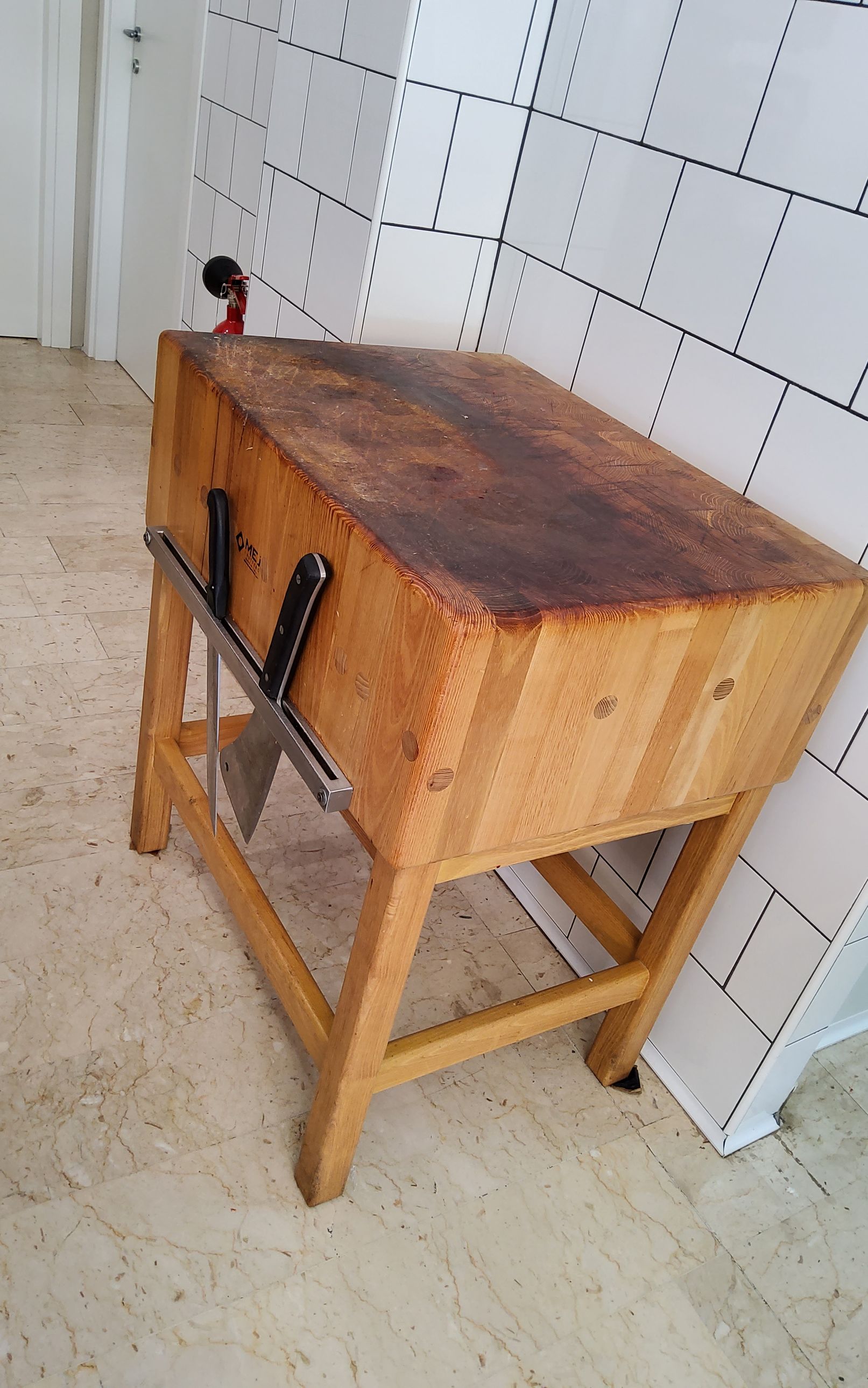 Ceppo da macellaio con due mannaie, appoggiato contro una parete piastrellata bianca. Il ceppo di legno è molto usato, con la parte superiore macchiata di marrone.