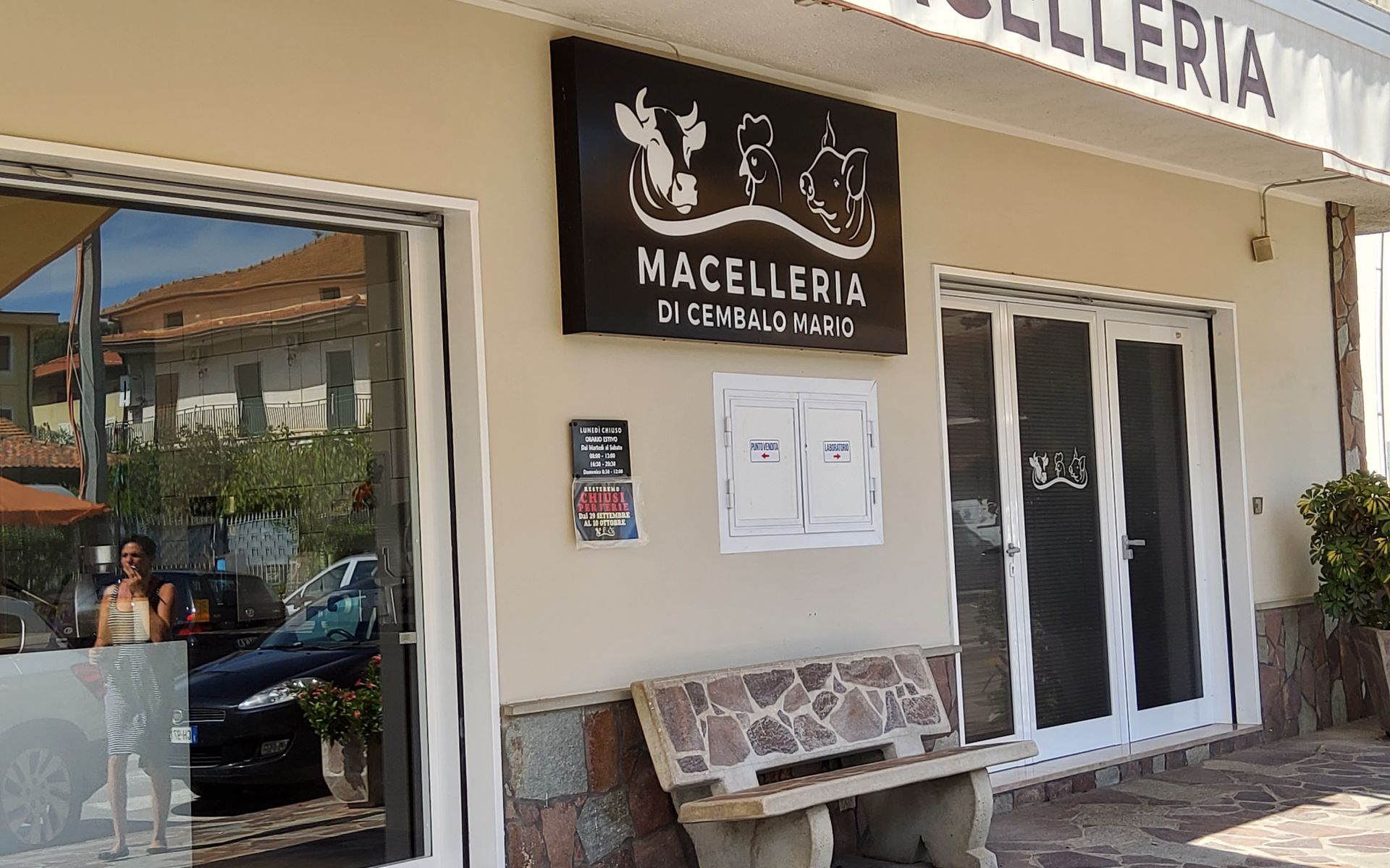 Esterno di una macelleria con insegna; panca in pietra; finestre in vetro.