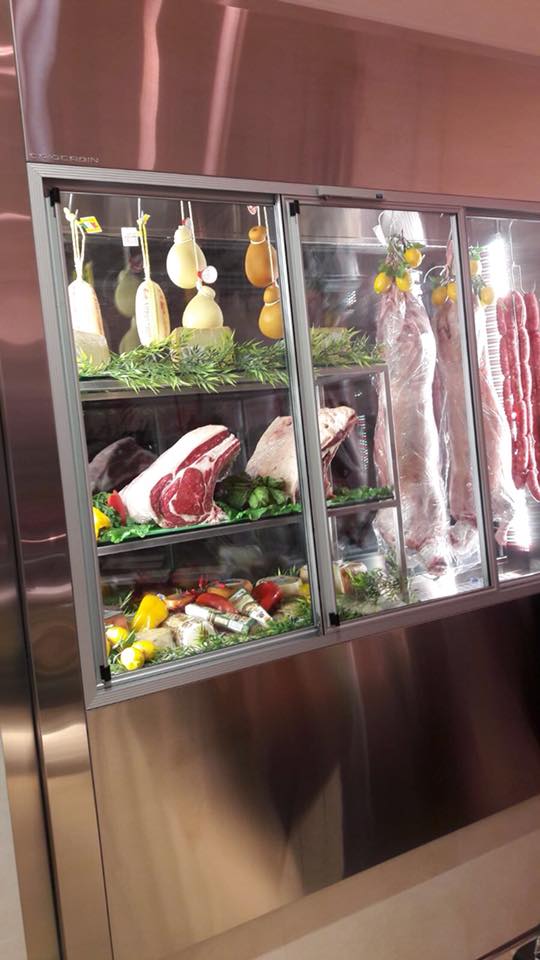 Vetrina refrigerata con salumi e formaggi vari, alcuni dei quali appesi, all'interno di un supporto in acciaio inox.