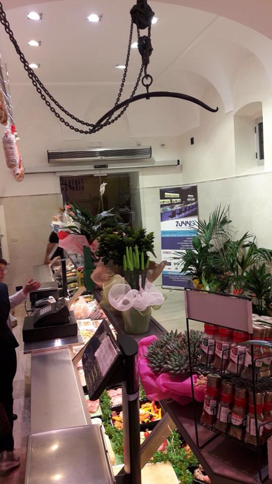 Interno di una macelleria con esposizione della carne, clienti e una bilancia sospesa.