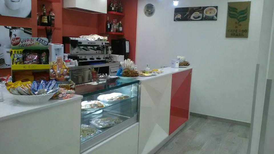 Interno di un bar con bancone con dolci, macchina per caffè espresso e scaffali con snack. Dettagli rossi e bianchi.