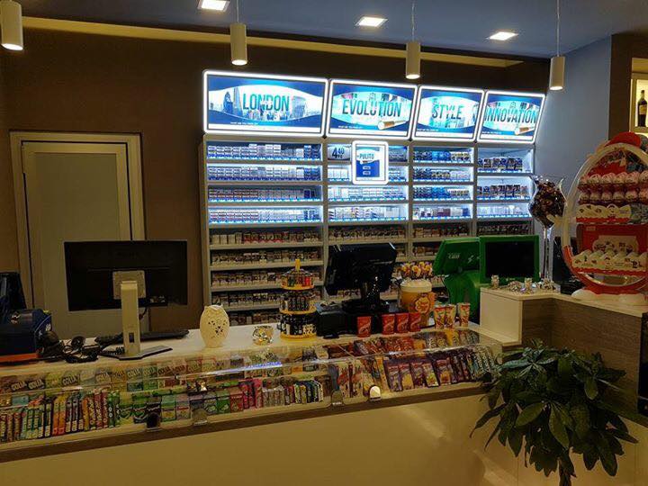 Interno di un minimarket, sigarette dietro il bancone, cassa, snack e una pianta.