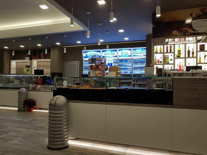 Vista interna di un bar con banconi espositivi, scaffali e un bar. Illuminazione dall'alto e pareti grigie.