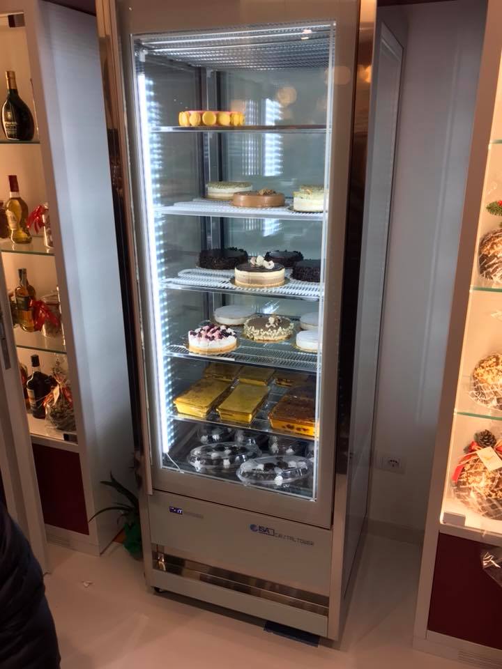 Vetrina refrigerata piena di pasticcini e torte, illuminata da luci interne.