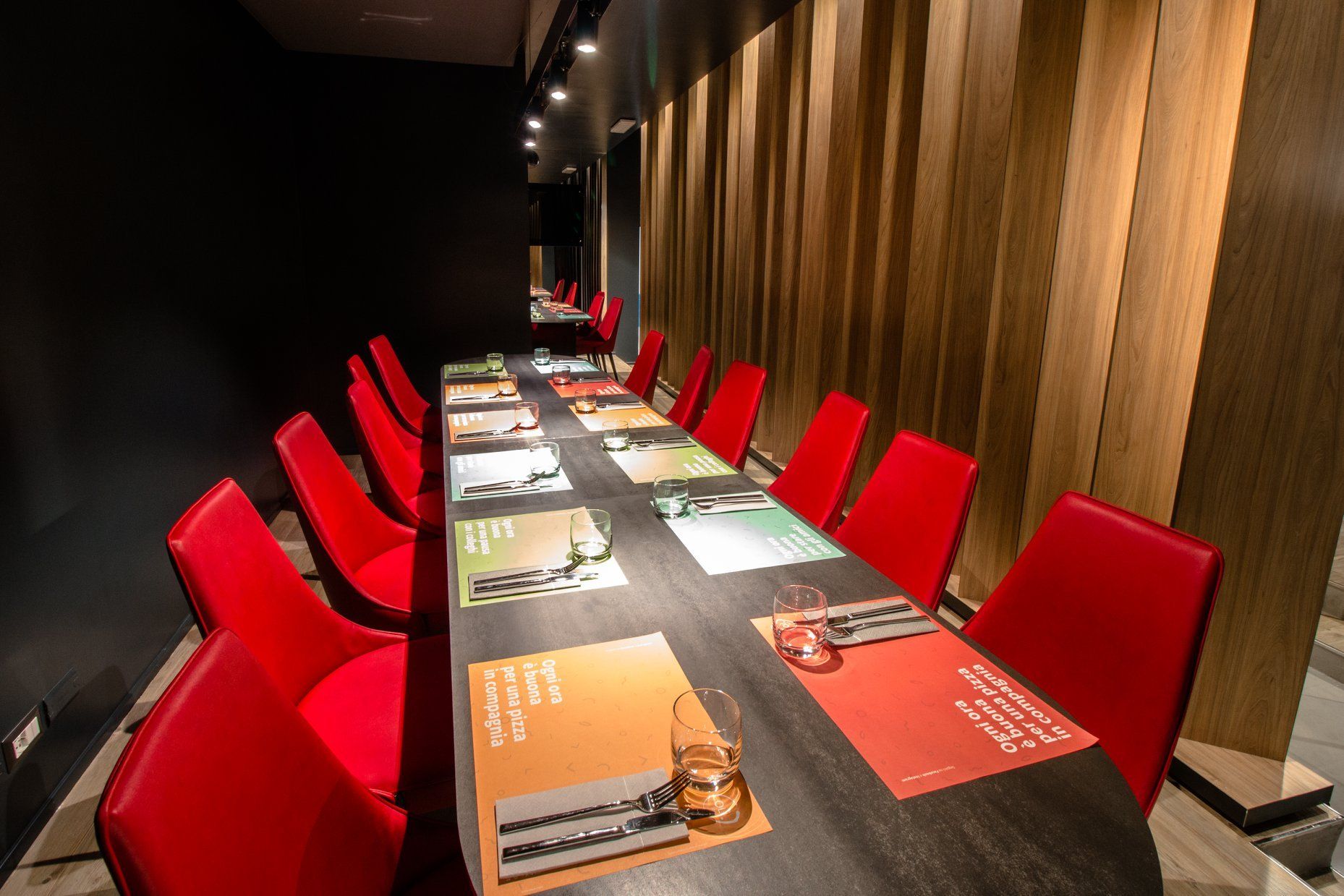 Un lungo tavolo in legno scuro apparecchiato con tovagliette all'americana e sedie rosse, in una moderna sala da pranzo allungata.