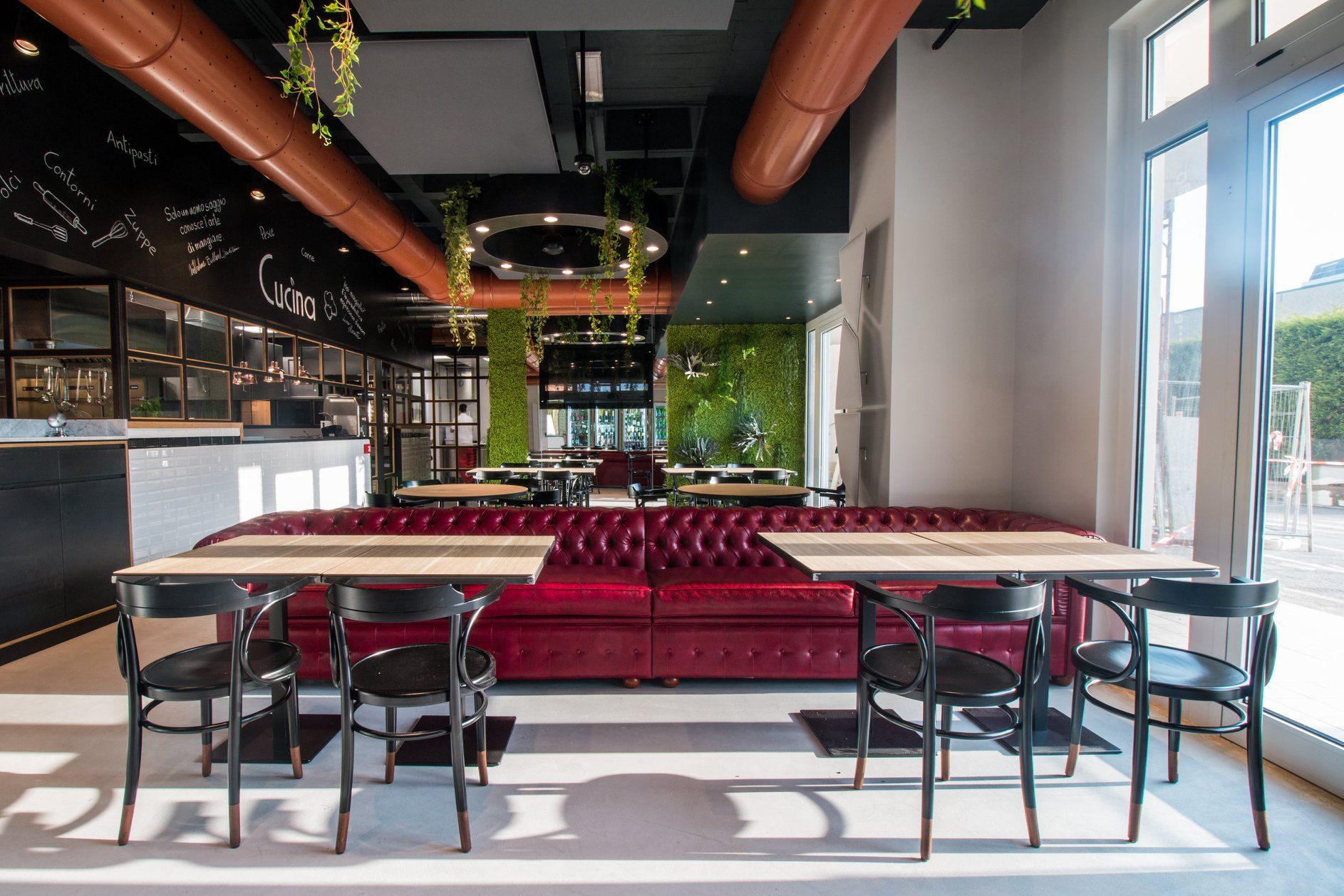 Interno del ristorante: cabina rossa con tavoli, sedie nere, piante e grandi finestre.