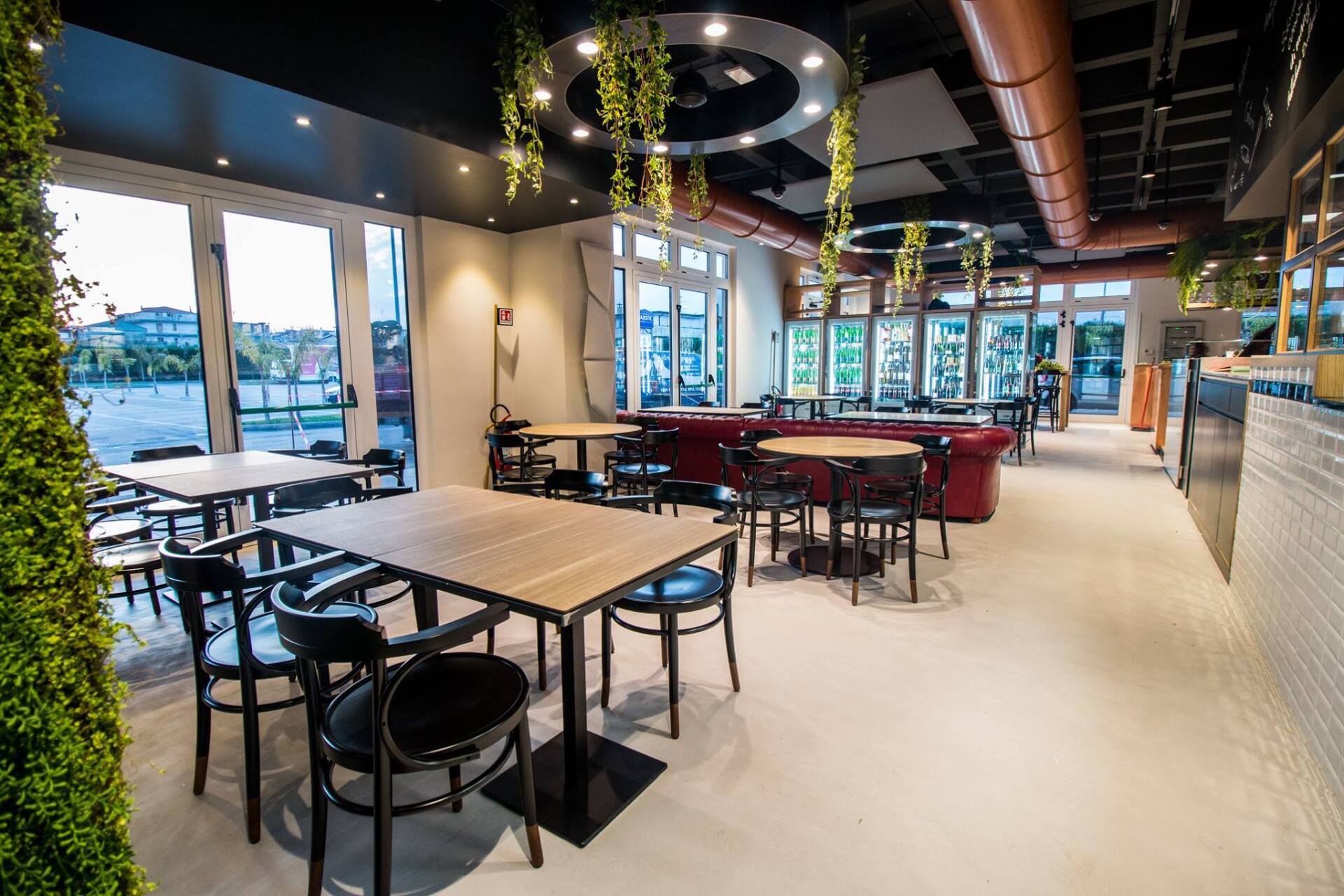 Interno del ristorante con tavoli e sedie, ampie finestre e vegetazione pensile.