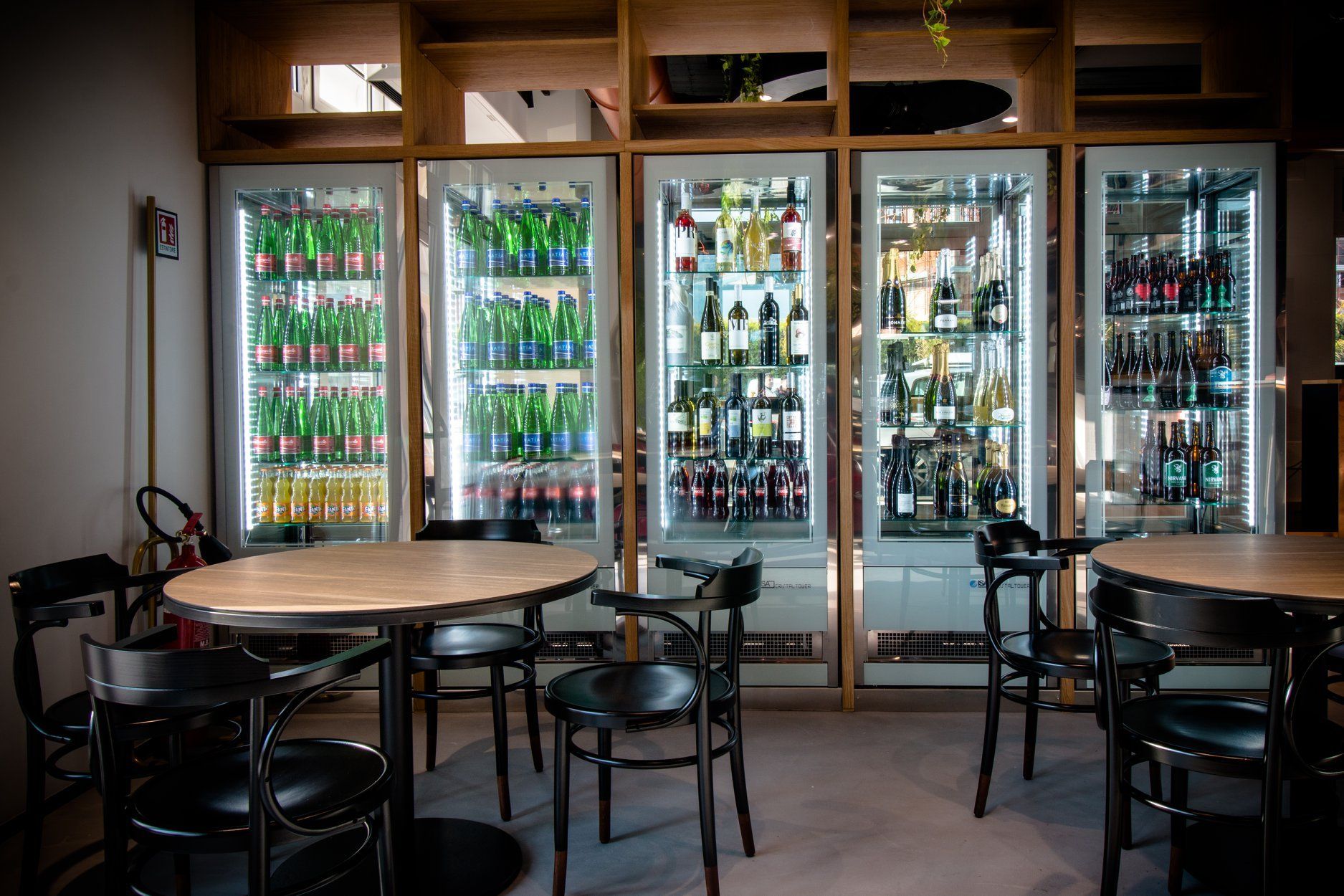 Interno del bar con tavoli rotondi, sedie nere e frigoriferi riforniti di bevande.