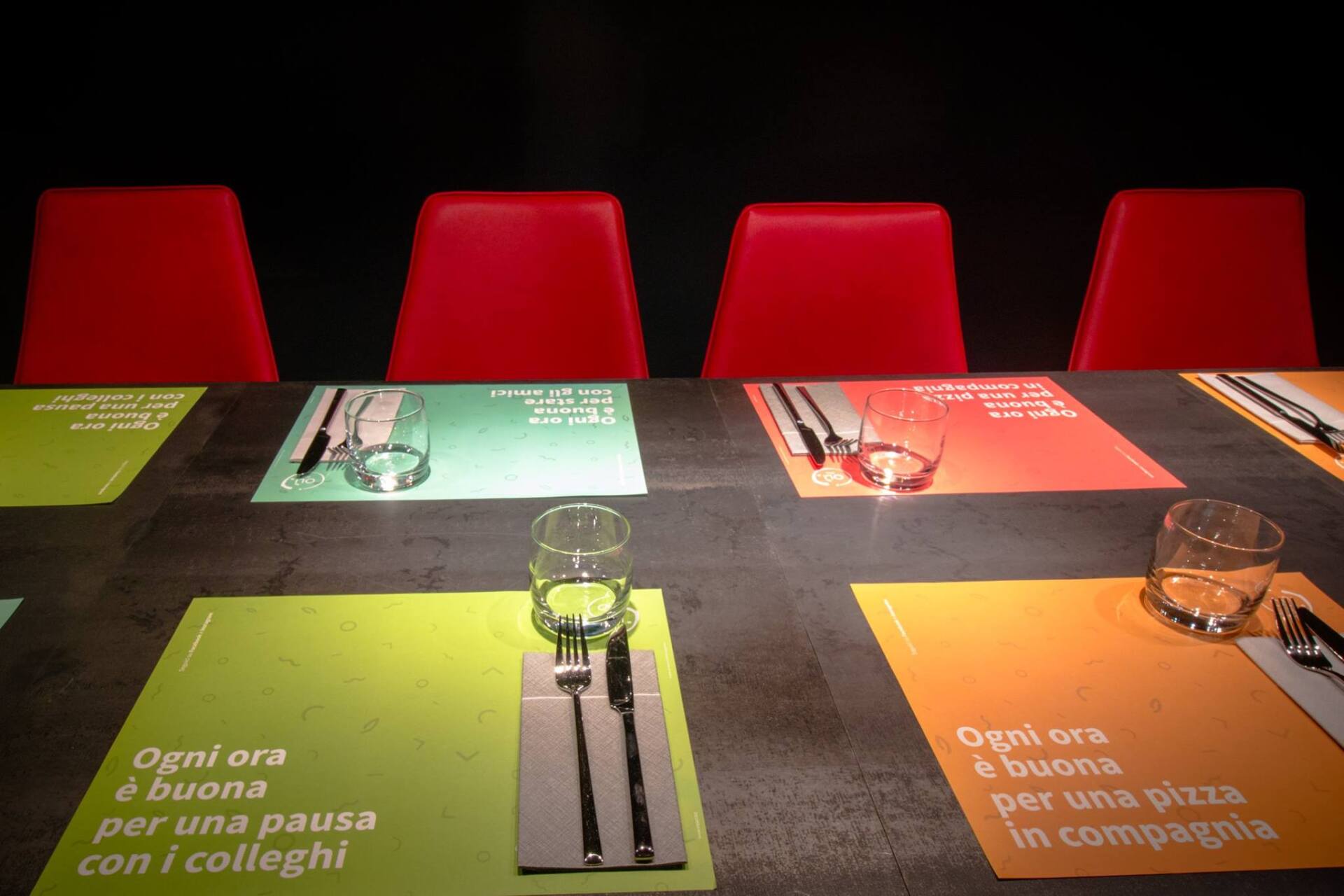 Tavolo del ristorante apparecchiato con tovagliette colorate, bicchieri, posate e sedie rosse su uno sfondo scuro.
