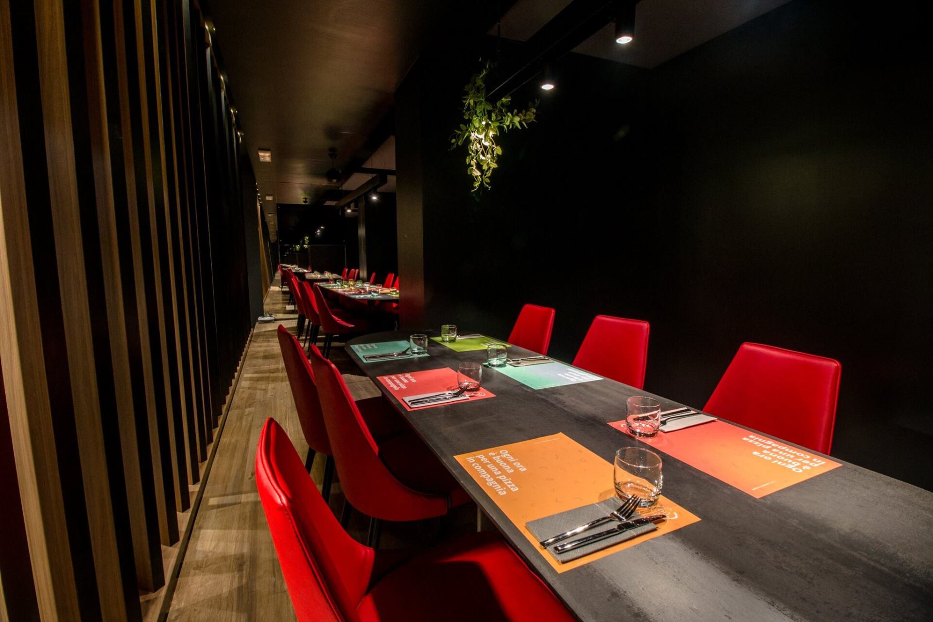 Lungo tavolo da pranzo rettangolare con sedie rosse e tovagliette colorate, in una stanza scarsamente illuminata.