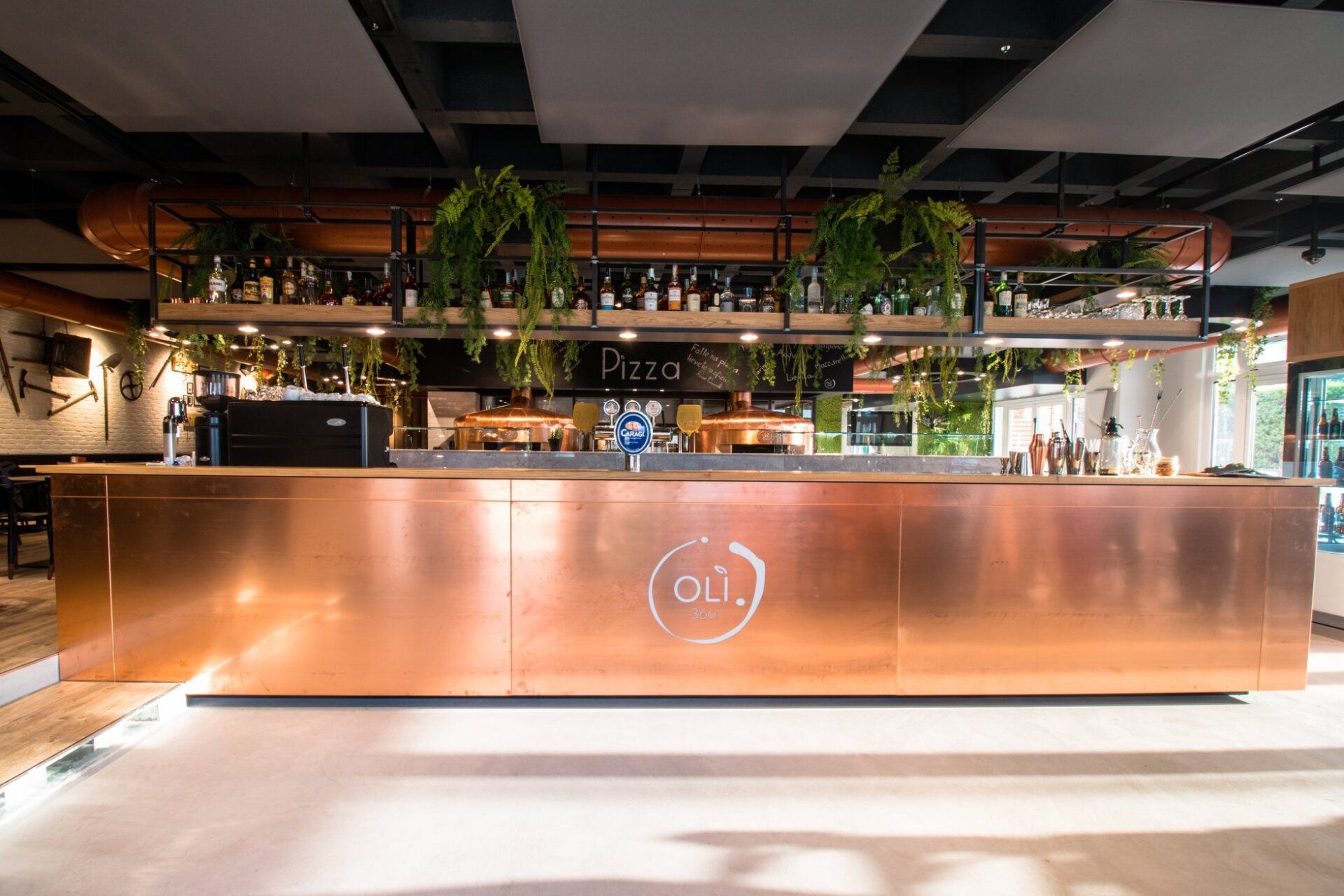 Bancone in rame con bottiglie, piante e logo, all'interno di un moderno ristorante.