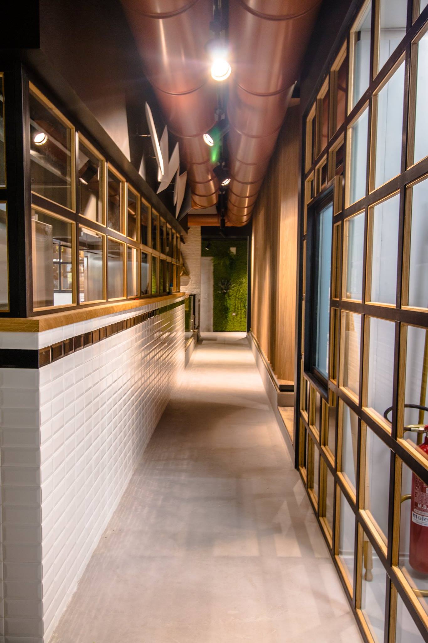 Corridoio stretto con muro di mattoni bianchi, pannelli di vetro e tubi di rame sopra la testa.