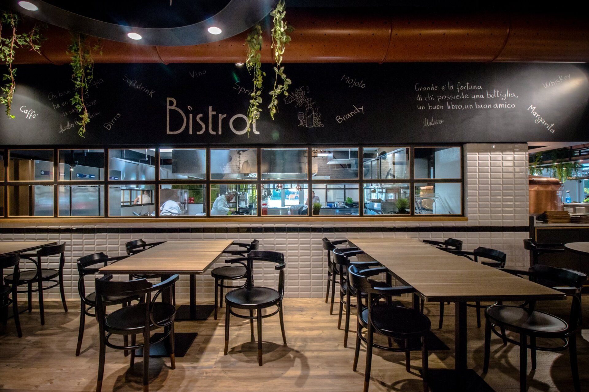 Interno del ristorante con tavoli, sedie e una finestra che si affaccia sulla cucina. La parola