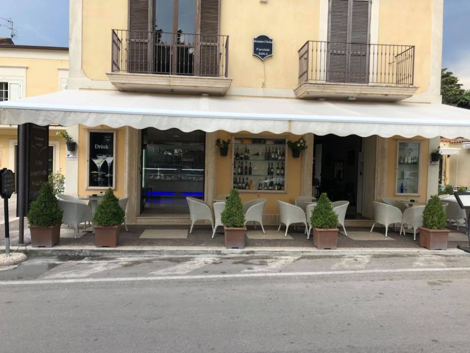 Esterno di un bar con una tenda bianca, alberi in vaso e posti a sedere all'aperto lungo una strada.