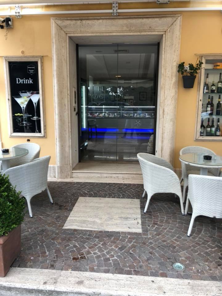 Esterno di un bar con porta a vetri, tavoli e sedie. È visibile l'insegna