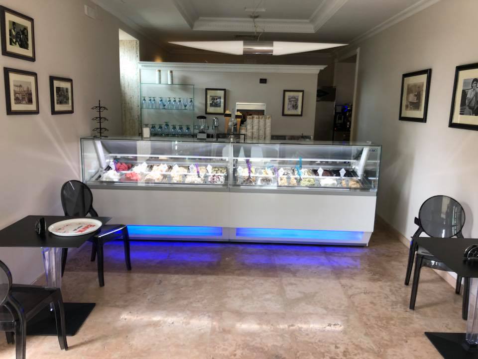 Interno di una gelateria con vetrina di gusti, tavoli, sedie e opere d'arte incorniciate.