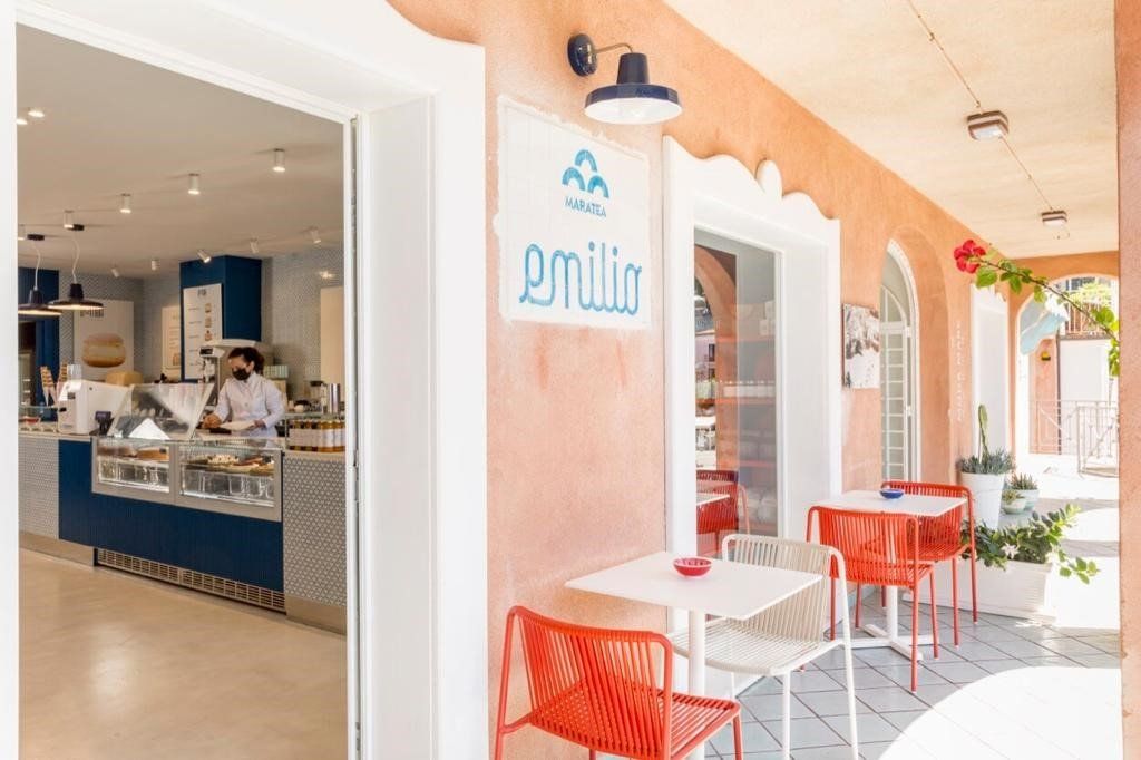 Esterno del bar Emilia con sedie e tavoli rossi; pareti color arancione pastello.