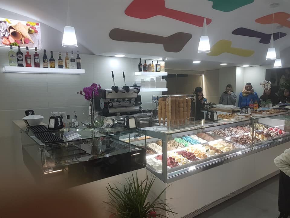 Interno di una gelateria con banco gelati, macchina per caffè espresso e clienti. Cucchiaini colorati decorano il soffitto.