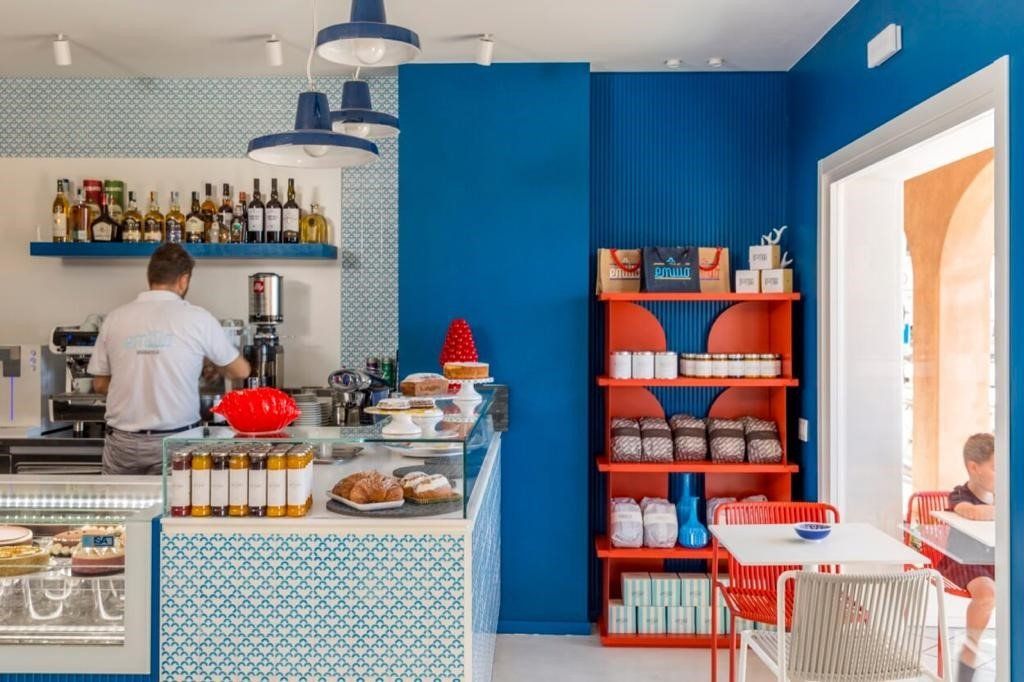 Interno di un bar con pareti blu, scaffali rossi, tavoli bianchi, un barista e pasticcini.