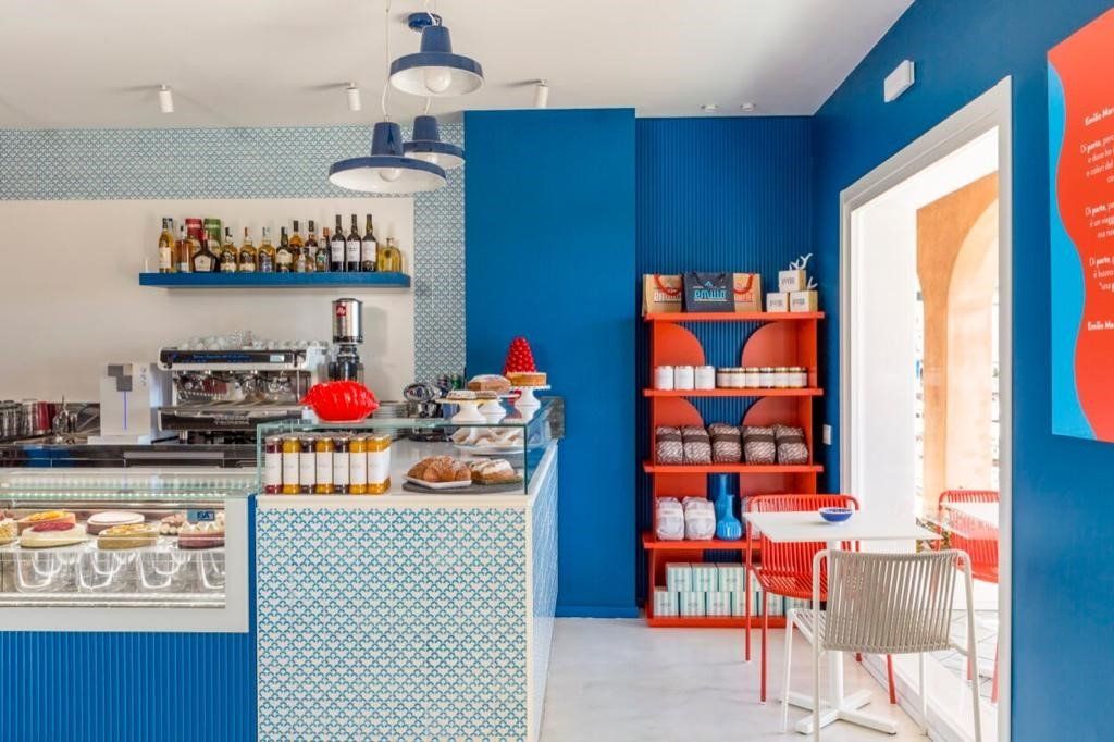 Interno del bar nei toni del blu e dell'arancione, con scaffali, bancone espositivo e tavoli.