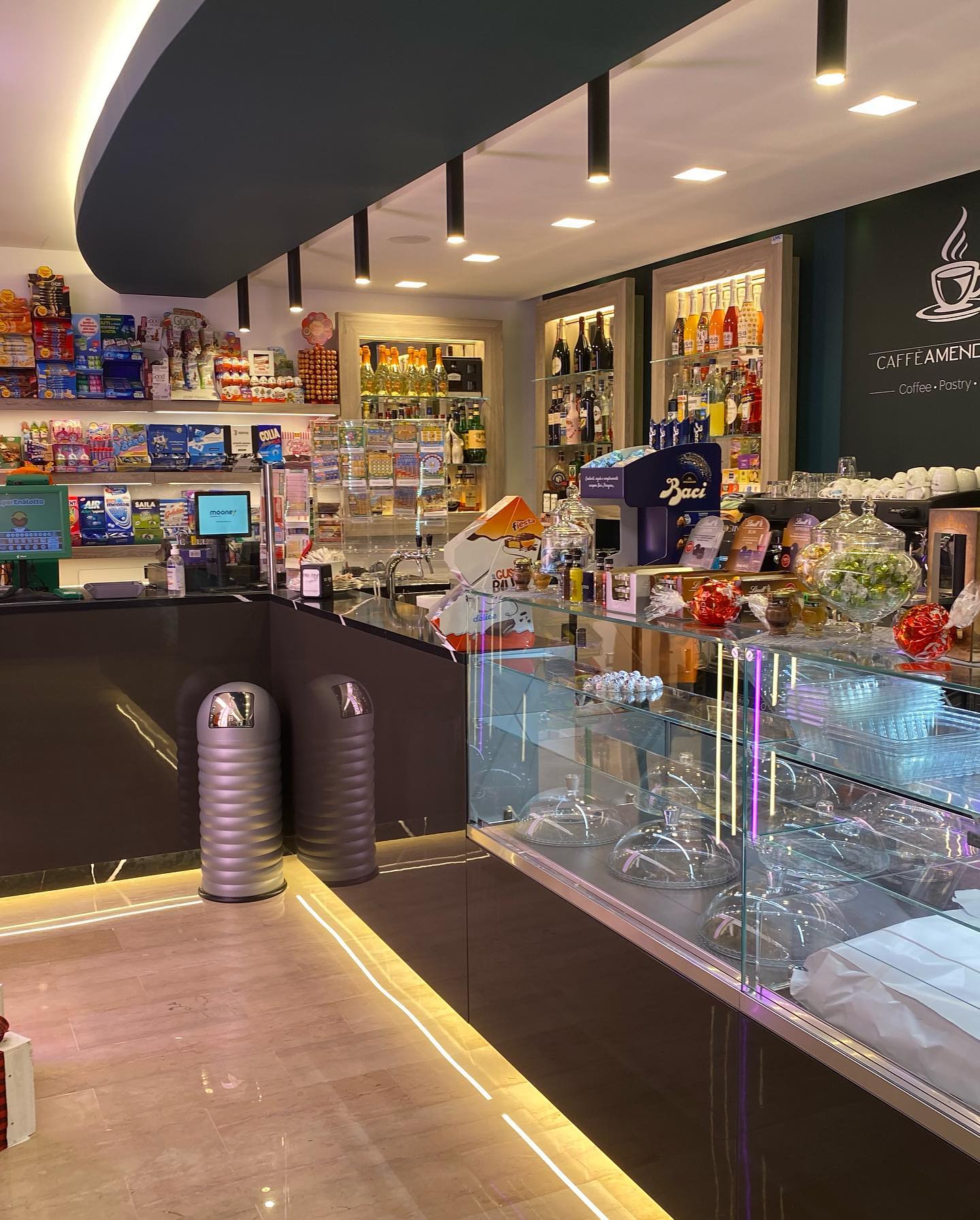 Interno di un bar con bancone, scaffali con prodotti, macchina per il caffè e area salotto.