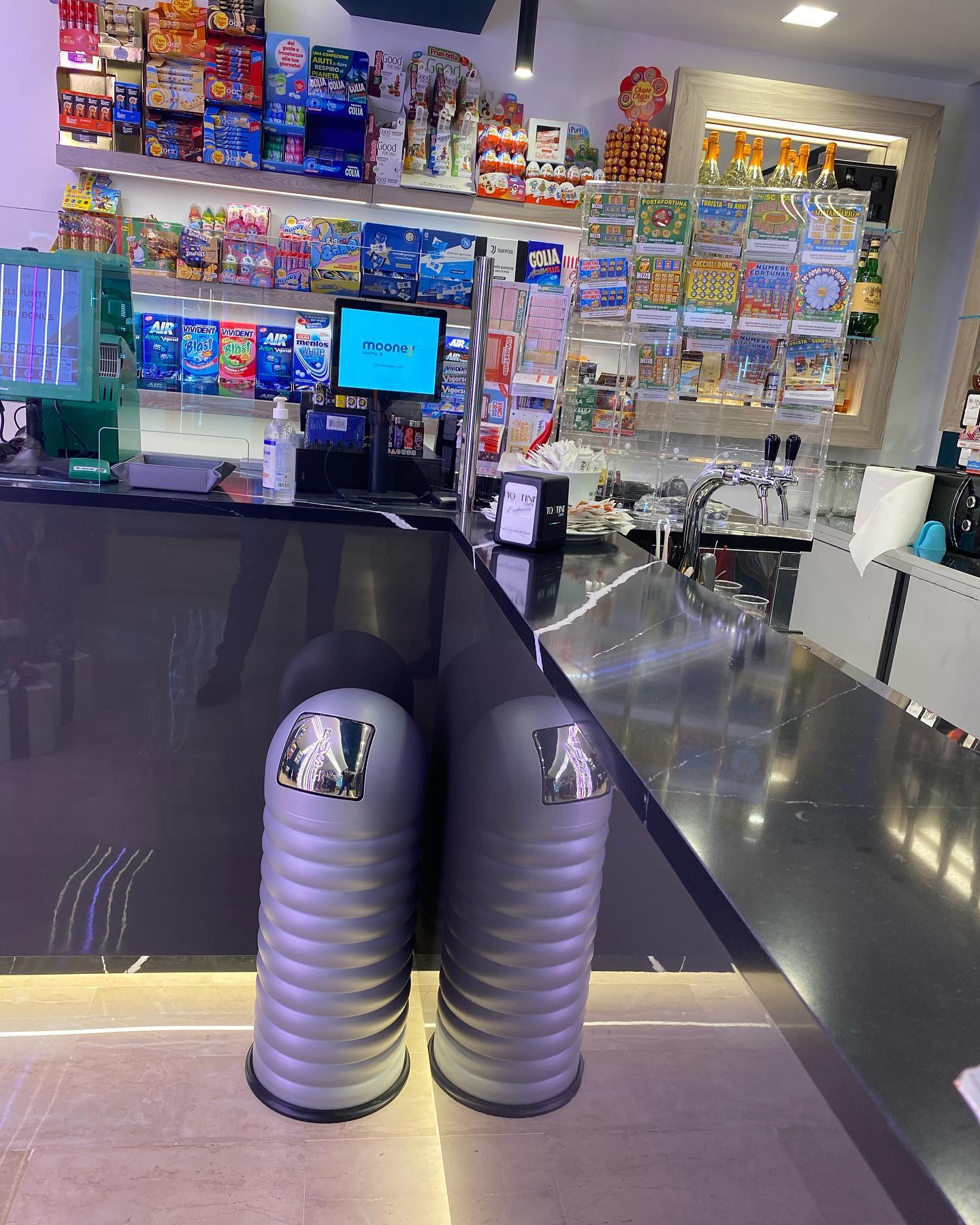 Bancone del bar con bevande e snack sugli scaffali di un negozio. Davanti a loro ci sono dei cestini grigi per la spazzatura.