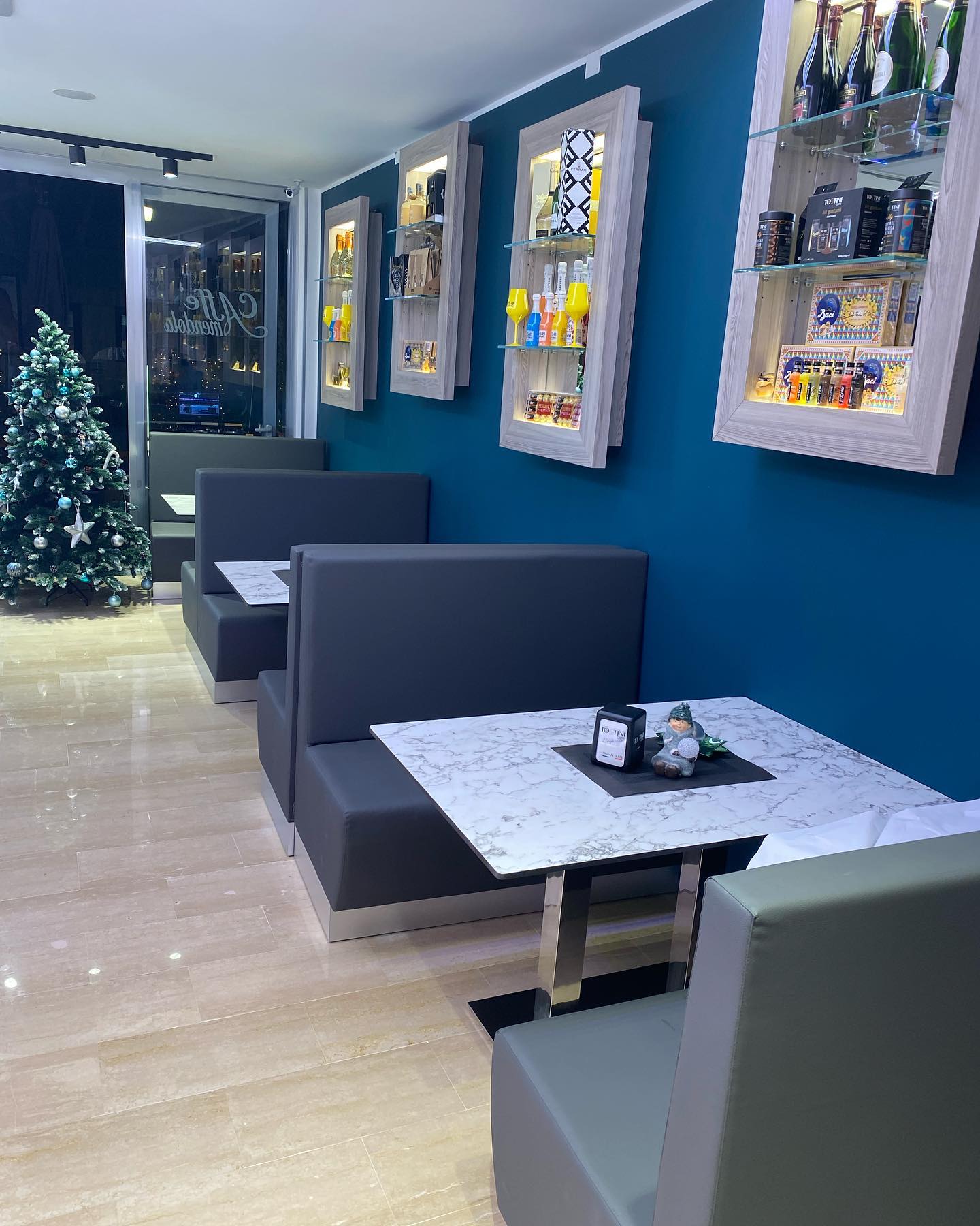 Interno di un bar con panche, tavoli e scaffali espositivi su una parete blu. Un albero di Natale si trova vicino a una finestra.