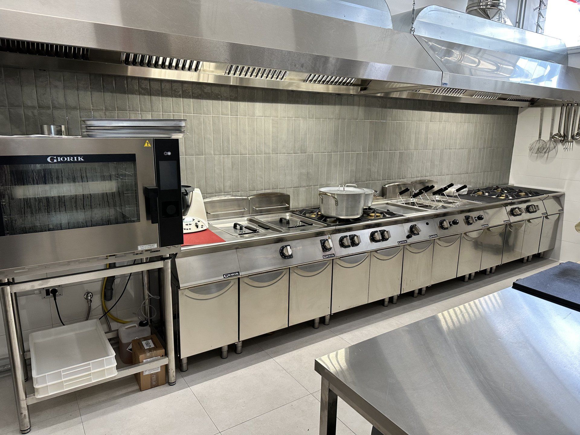 Cucina commerciale con elettrodomestici in acciaio inossidabile, tra cui forno, fornelli e sistema di ventilazione.