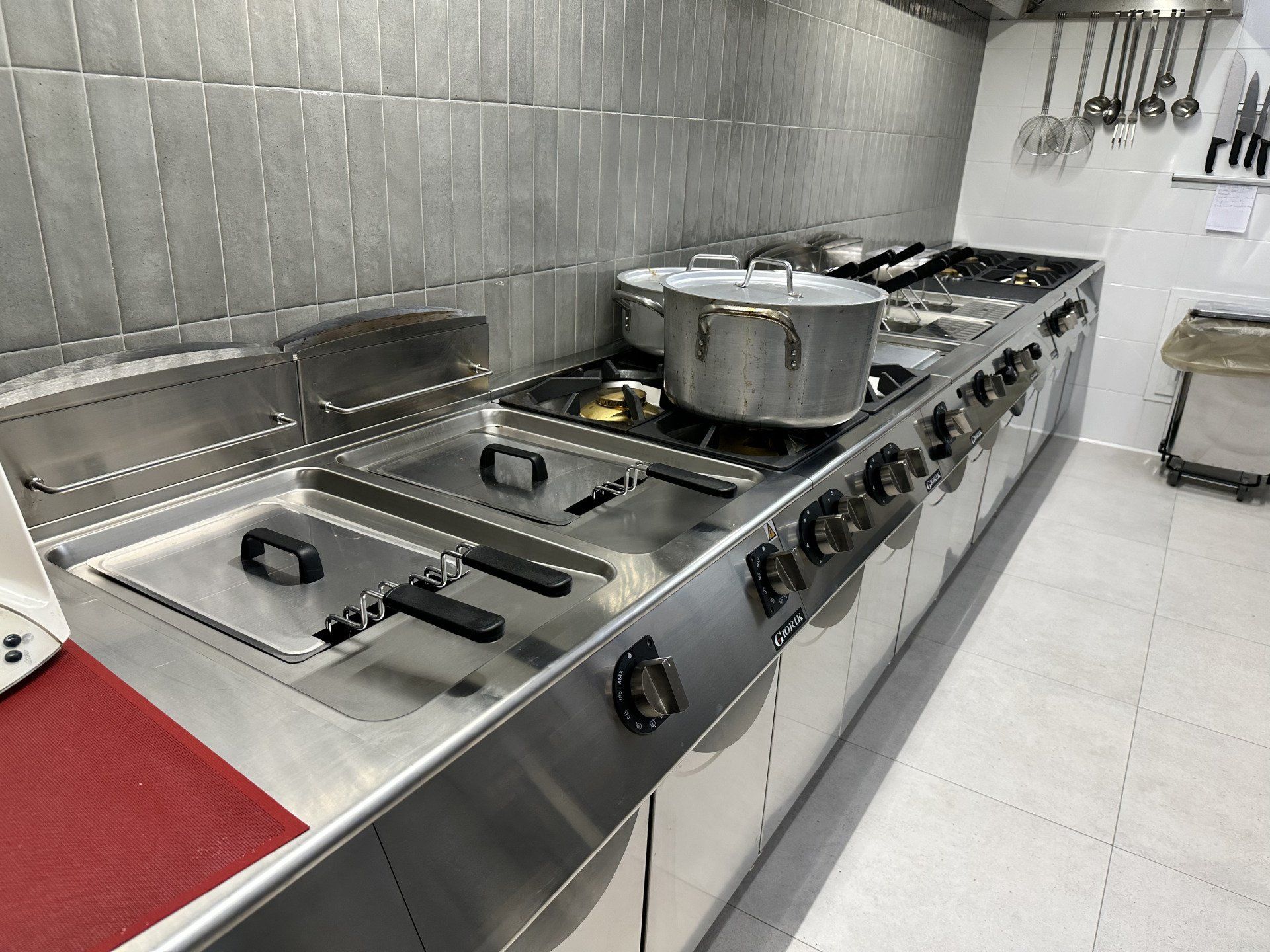 Cucina commerciale con pentole e friggitrici, acciaio inossidabile, paraschizzi in piastrelle.