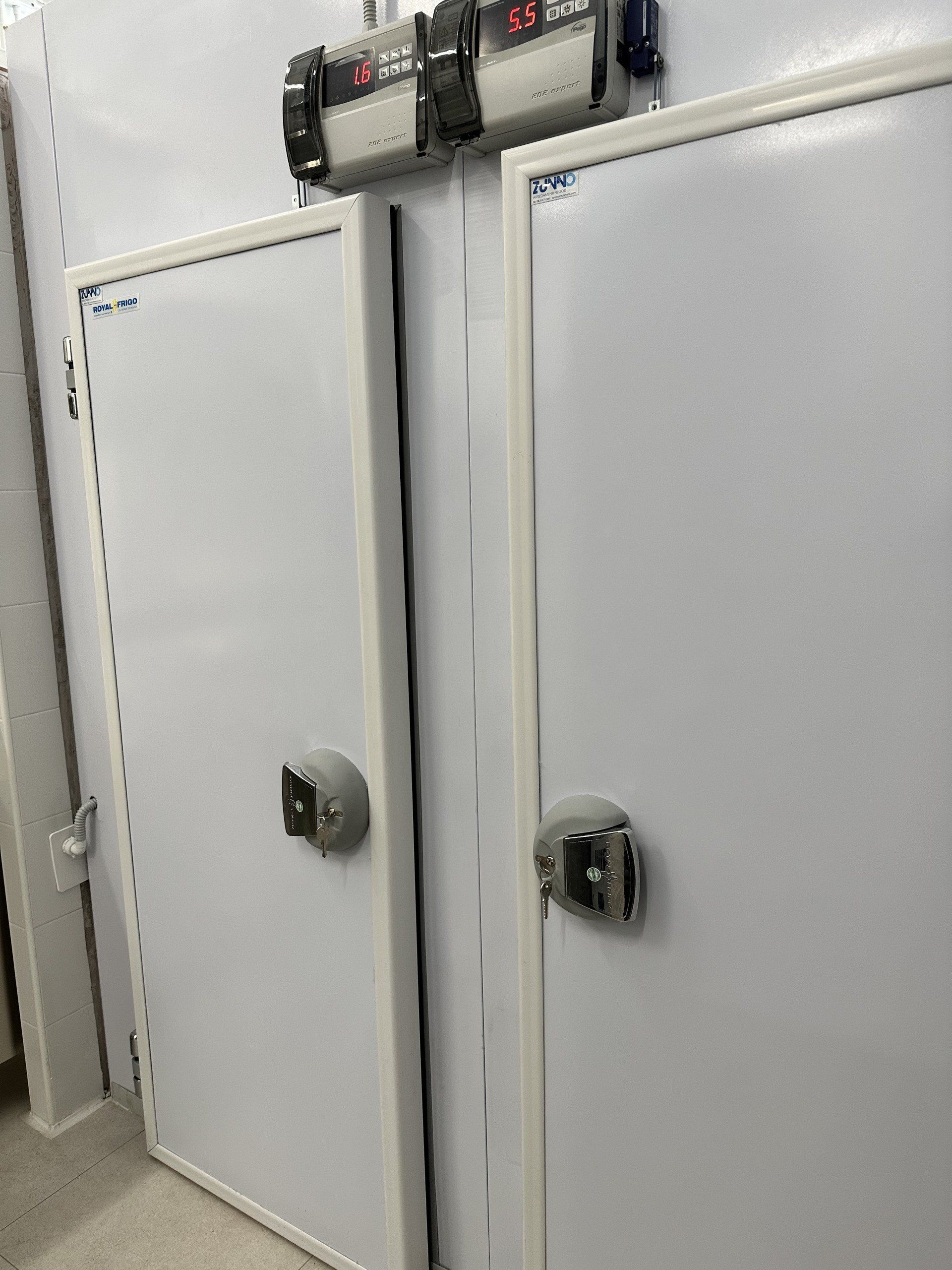 Due porte bianche di una cella frigorifera con chiavistelli, fiancheggiate da pareti bianche. In alto si trovano i comandi.