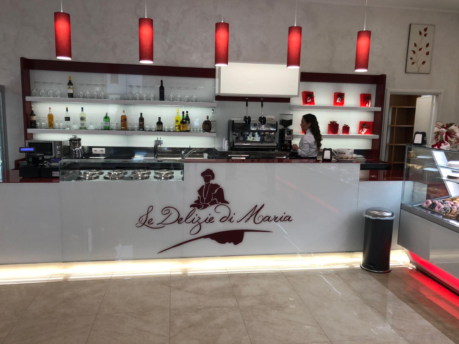 Interno del bar con una donna dietro al bancone, bottiglie e una macchina per il caffè. Logo