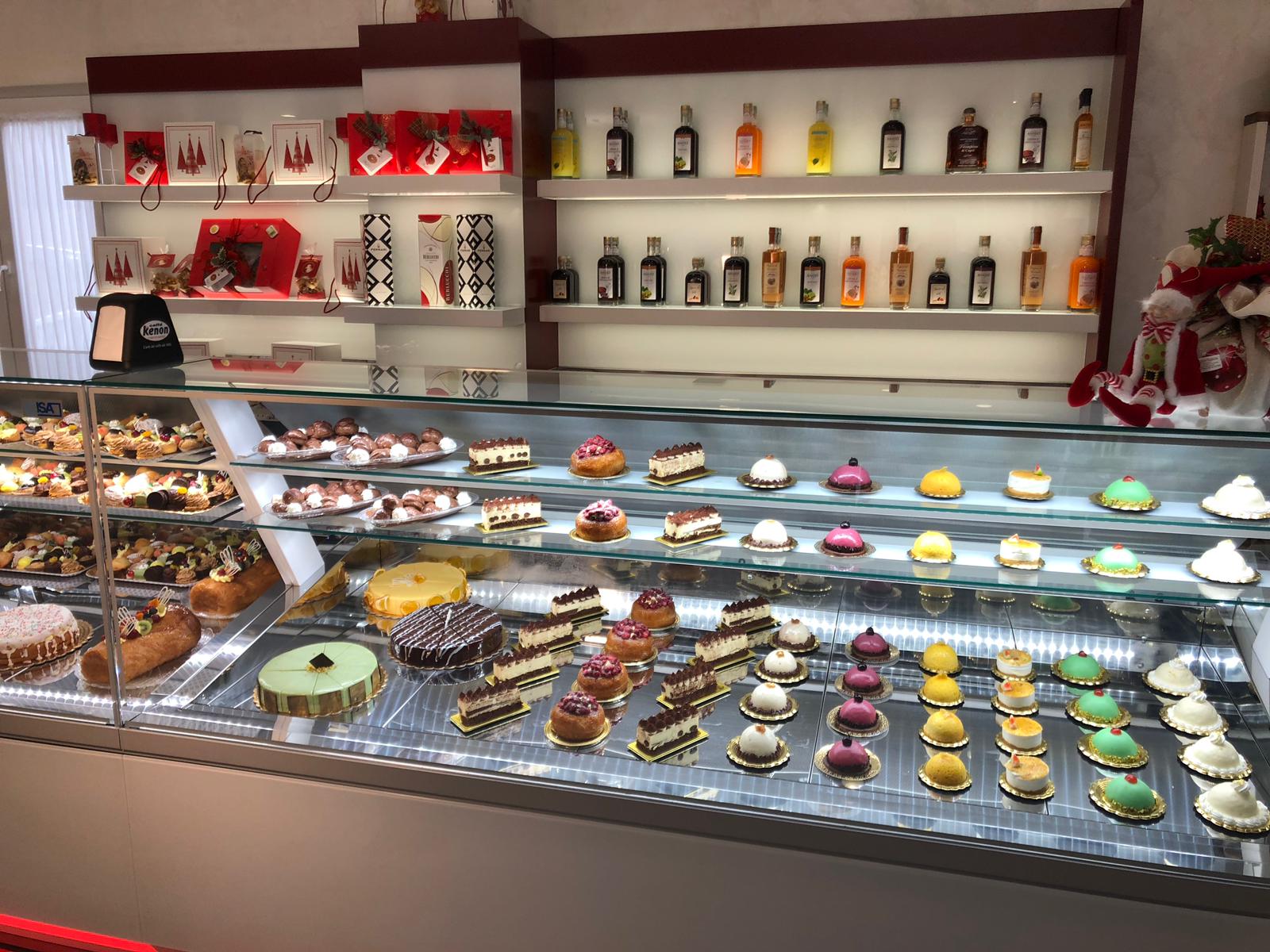 Vetrina di pasticceria in un negozio pieno di torte e dessert; sullo sfondo scaffali con bottiglie e scatole.