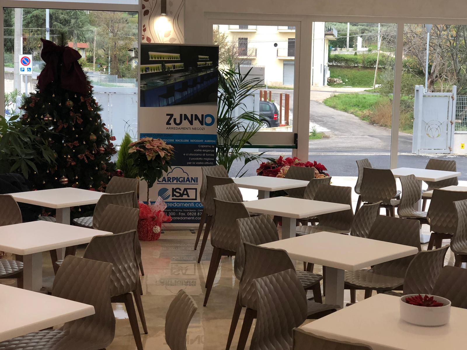 Mensa con tavoli bianchi e sedie grigie. Decorazioni natalizie e striscioni visibili; vista attraverso le finestre.