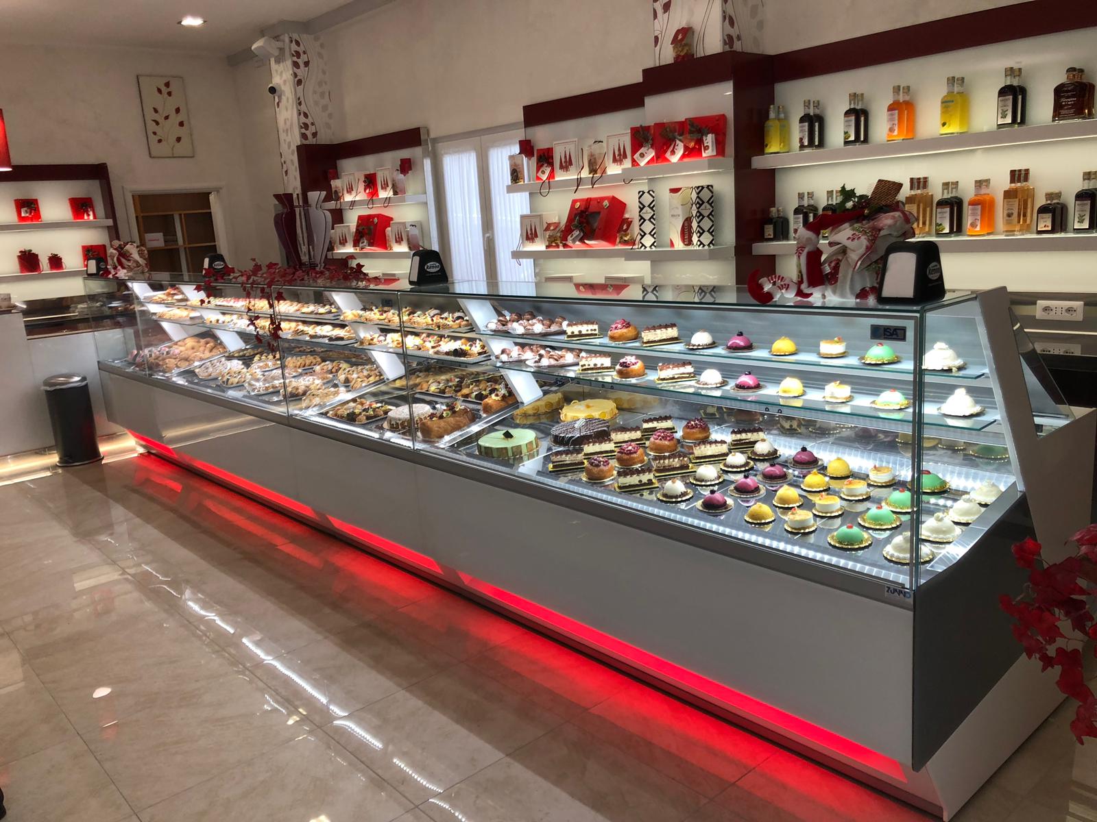 Una vetrina da panetteria illuminata, piena di pasticcini e torte. Luci d'accento rosse.