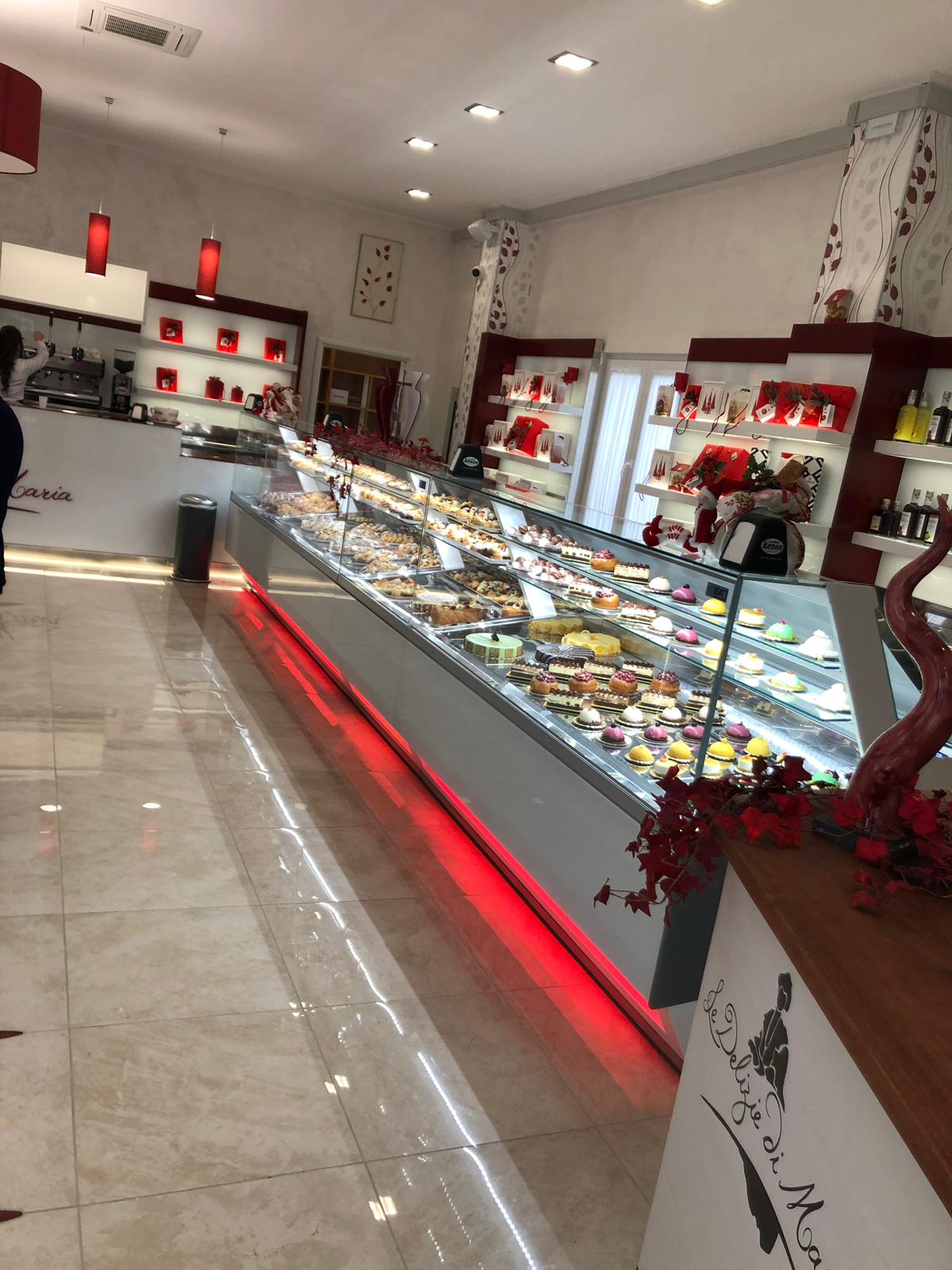Interno di una pasticceria con una lunga vetrina piena di pasticcini, illuminazione rossa e decorazioni.