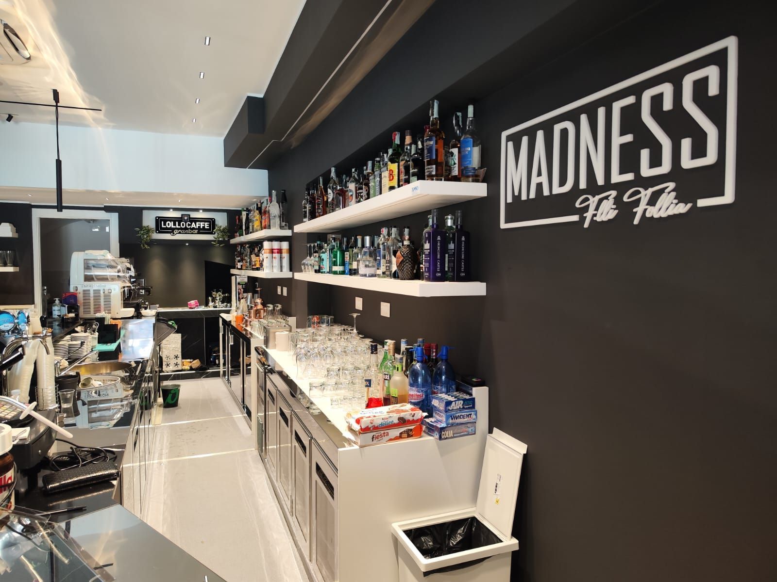 Interno del bar con scaffali pieni di liquori, bicchieri e un cartello con la scritta