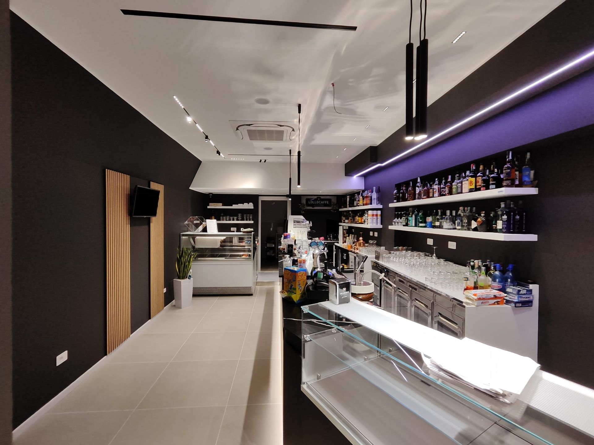 Interno di un piccolo bar, con un bancone con bottiglie, vetrine e un lungo corridoio con pareti scure e illuminazione moderna.