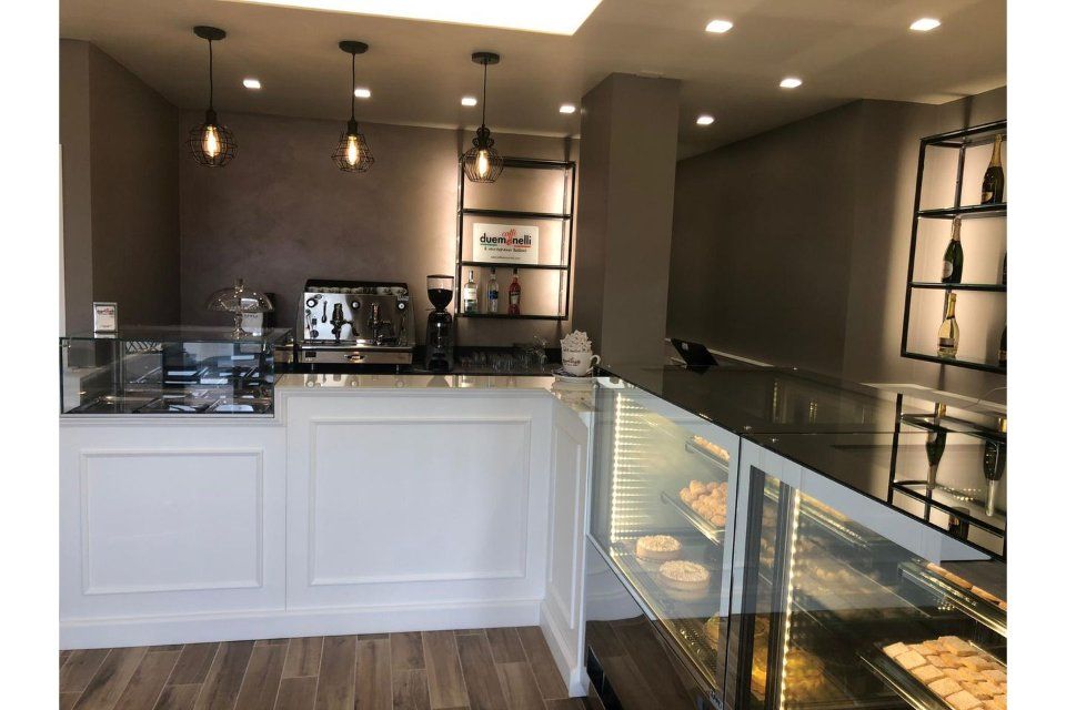 Interno di un bar con bancone bianco, vetrina per dolci, macchina per l'espresso e luci sospese.