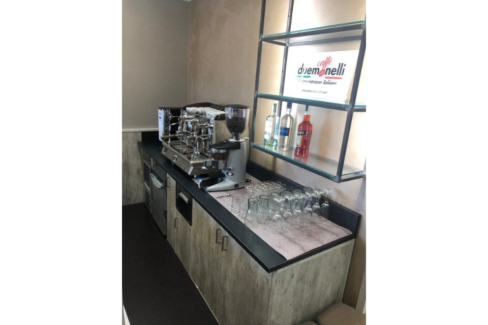 Macchina per caffè espresso e allestimento bar in uno spazio commerciale, con scaffali, bottiglie di liquore e bicchieri.