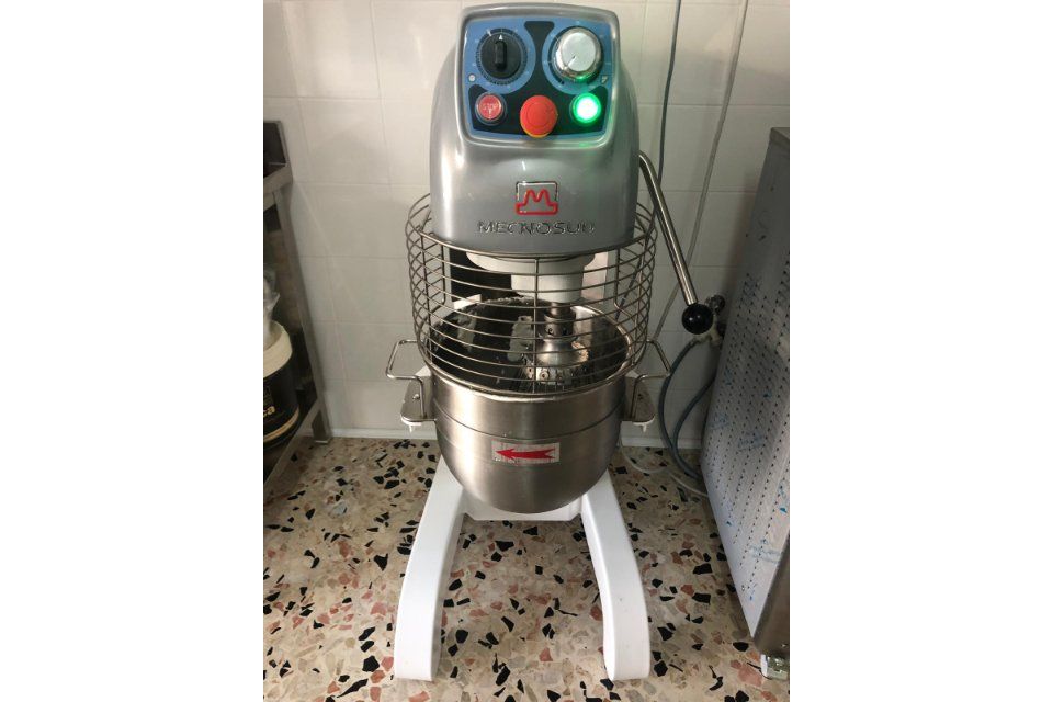 Un robot da cucina professionale con ciotola in acciaio inox e gabbia protettiva. È argentato e bianco, su un pavimento decorato.