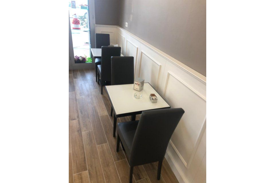 Posti a sedere all'interno del bar: tavoli quadrati con sedie nere, contro una parete bianca. Pavimento in legno.