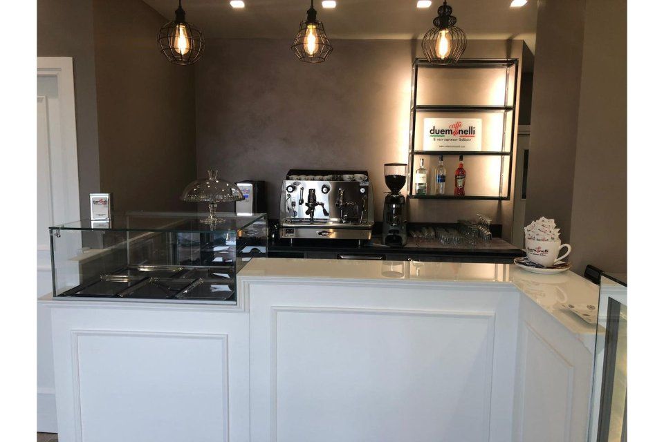 Bar con macchina per caffè espresso, macinacaffè e scaffali con prodotti; bancone bianco e pareti scure.